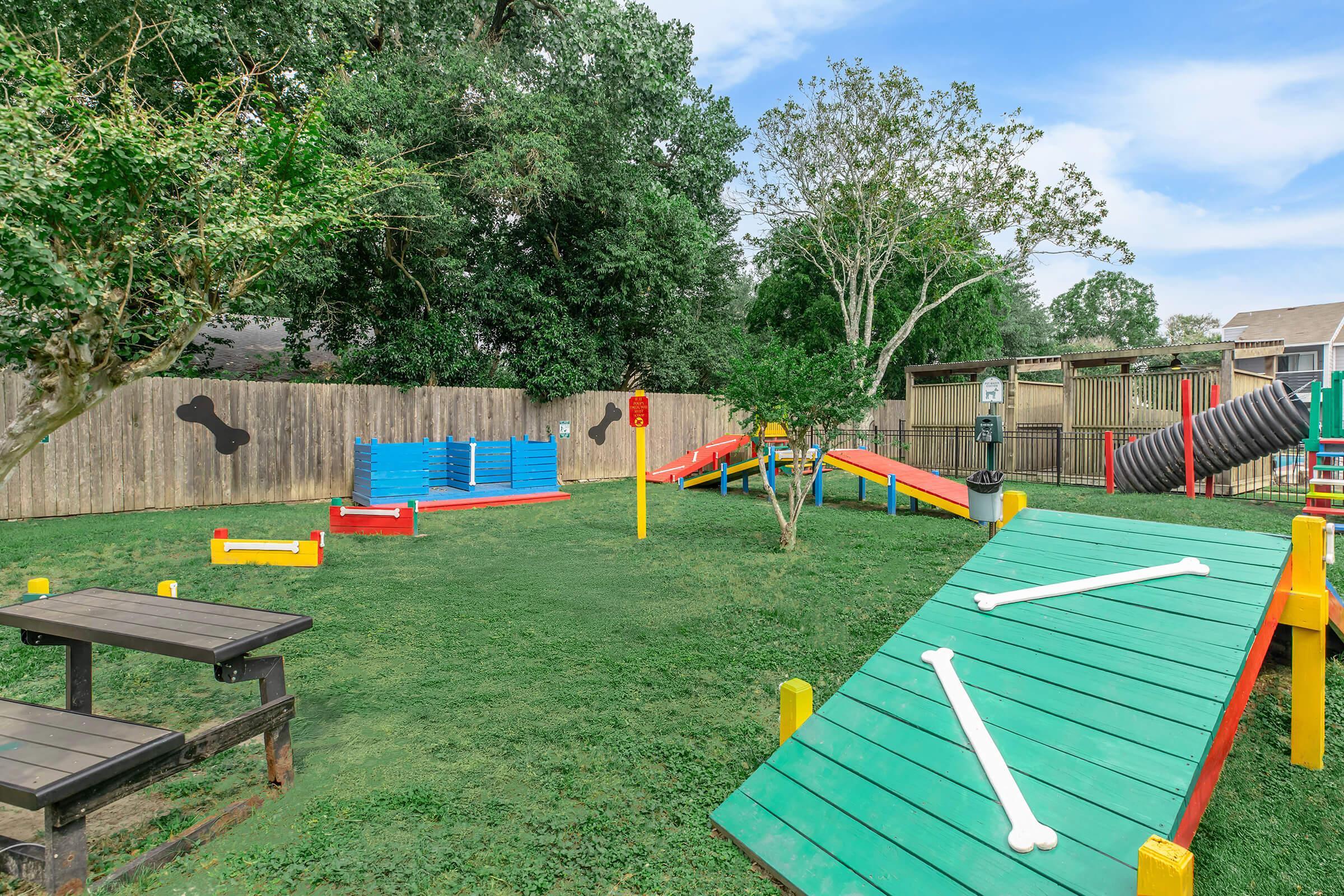 A vibrant dog park with green grass, colorful play equipment, and ramps for dogs. The area features benches and wooden structures, with a fenced background of trees and a clear blue sky. Brightly colored elements include large, oversized bone shapes on the ground, enhancing the playful atmosphere.