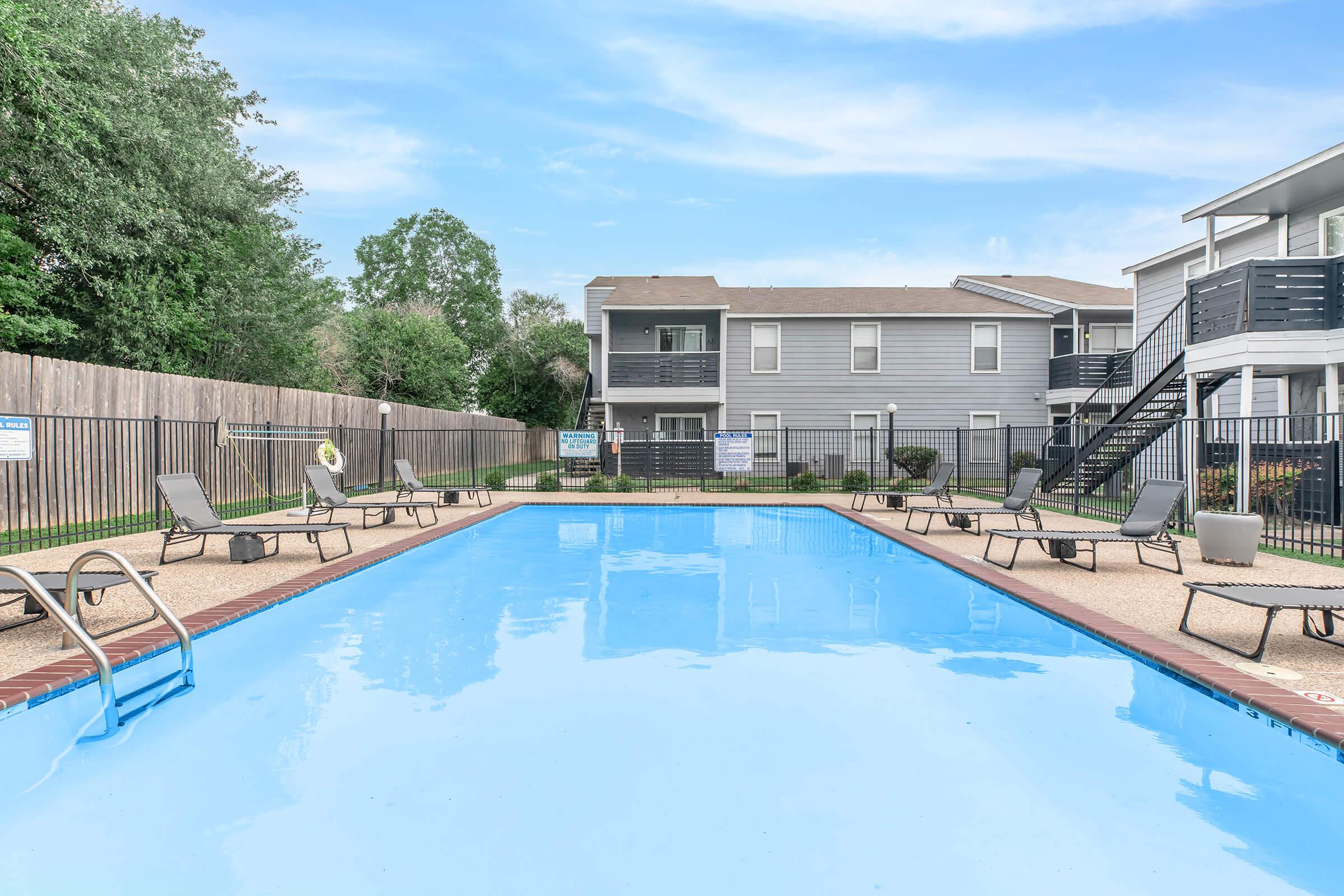 A swimming pool surrounded by lounge chairs and a fenced area, with an apartment building in the background. Lush greenery can be seen on the sides, and there's a staircase leading to the upper level of the building. The sky is clear and blue.