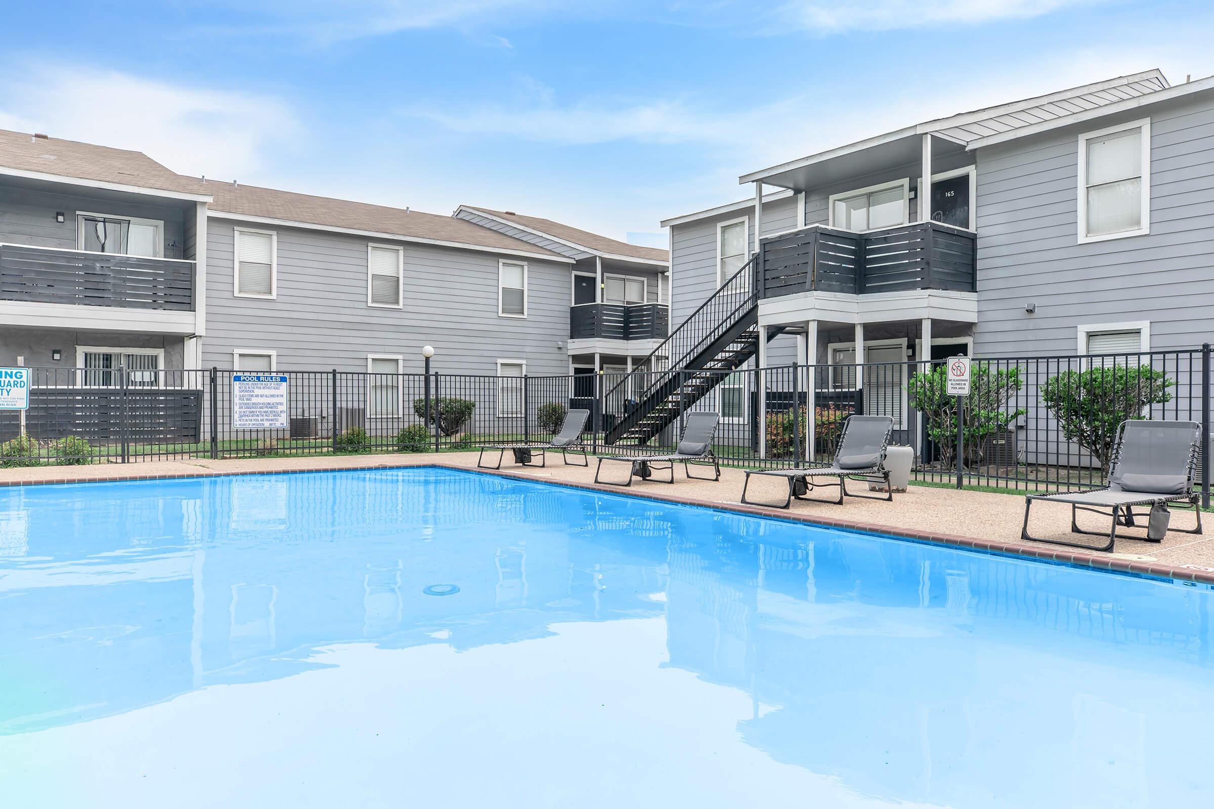 A clear blue swimming pool surrounded by lounge chairs, with gray apartment buildings in the background. The area features a fenced perimeter, and there are signs nearby. The sky is partly cloudy, creating a bright and inviting atmosphere.