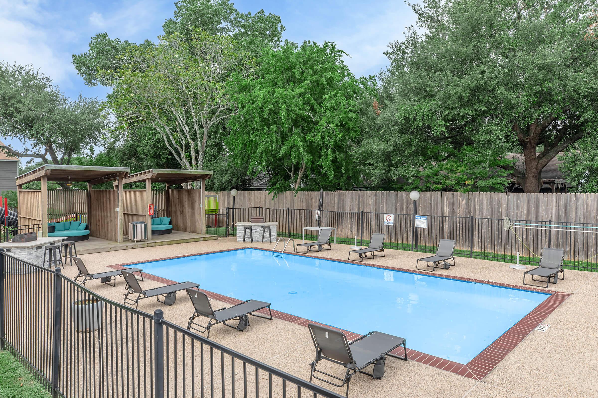 A serene outdoor swimming pool surrounded by lounge chairs and small cabanas. The pool area is enclosed by a fence, with trees and greenery in the background offering a relaxing atmosphere. The sky is partly cloudy, enhancing the bright and inviting setting.