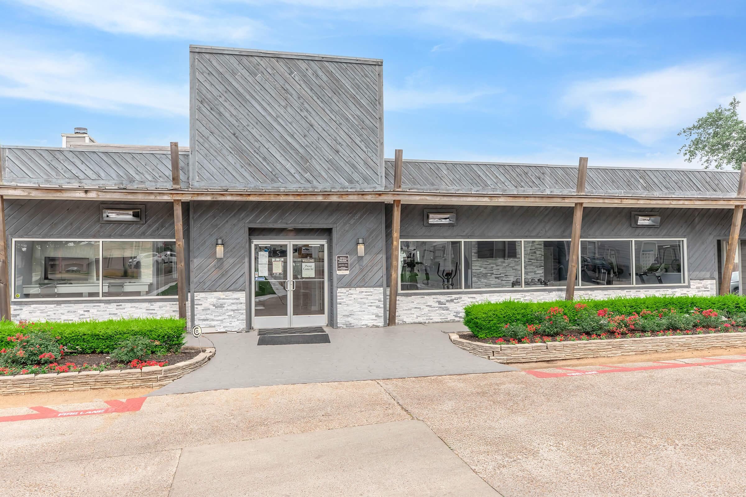 A modern one-story building with a gray exterior, featuring large windows, stone detailing at the base, and a flat roof. The entrance has glass doors and is flanked by landscaping with shrubs and flowers. The overall appearance is clean and contemporary, set against a clear blue sky.