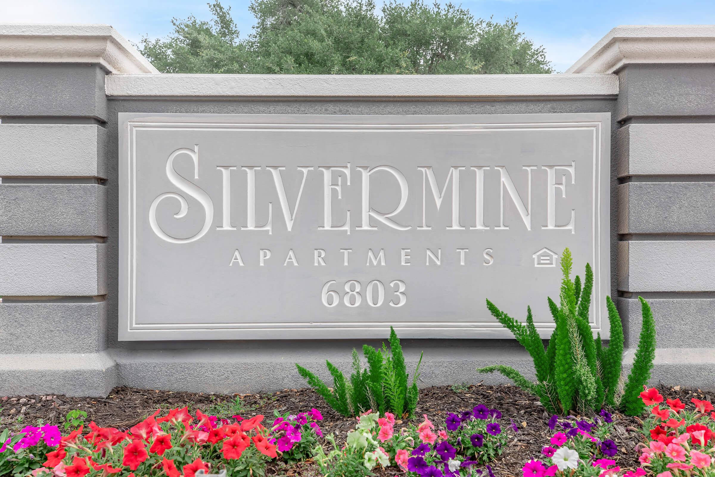 Sign for Silvermine Apartments, featuring the name "Silvermine" prominently displayed in elegant lettering, with the apartment number 6803 below. Surrounding the sign, there are vibrant flower beds with red, pink, and purple flowers, alongside green plants. The background shows trees and a clear sky.