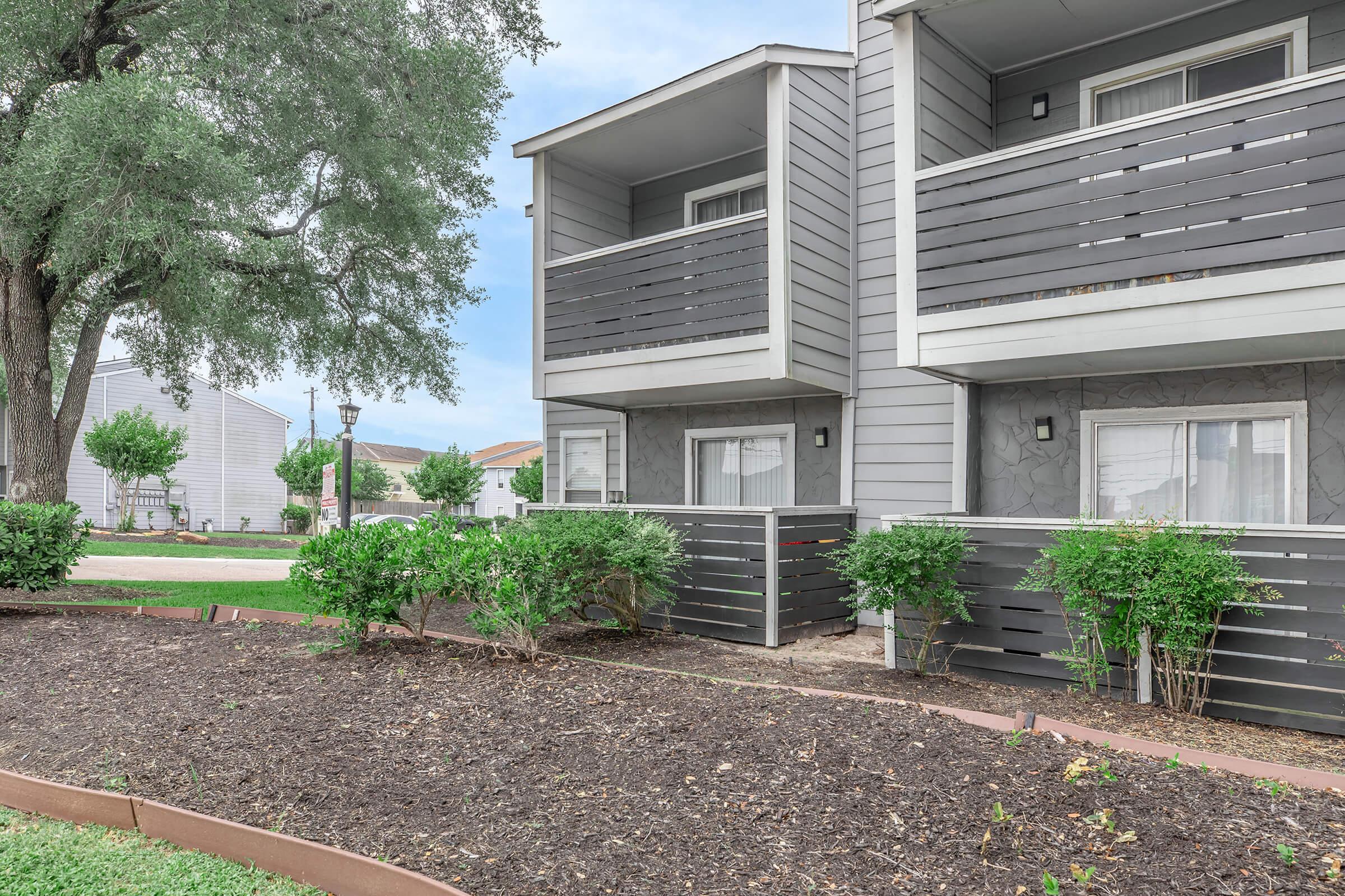 A view of a two-story residential building featuring balconies on the upper level. The exterior is clad in gray siding, with green shrubs planted in front. The surrounding area includes grass and landscaping, giving a well-maintained appearance. Blue sky is visible in the background.