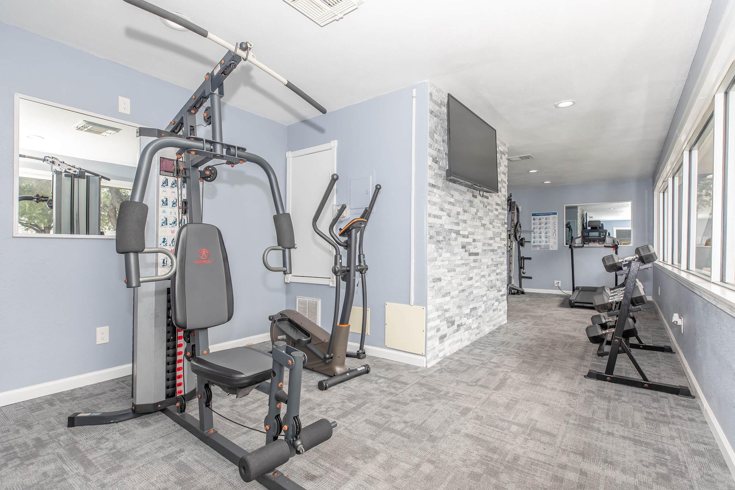 A modern gym interior featuring exercise equipment, including a multi-gym, an elliptical machine, and several weight benches. The walls are painted blue, and there are large windows allowing natural light in, along with a wall-mounted television and a mirror reflecting the workout area.
