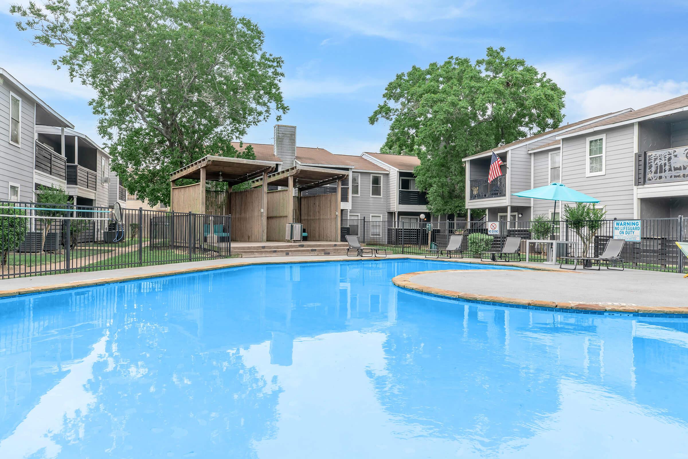 A swimming pool surrounded by a fence, with lounge chairs and an umbrella nearby. In the background, there are two multi-story apartment buildings and a shaded wooden structure. Green trees offer a natural ambiance, and the sky is clear with a few clouds.