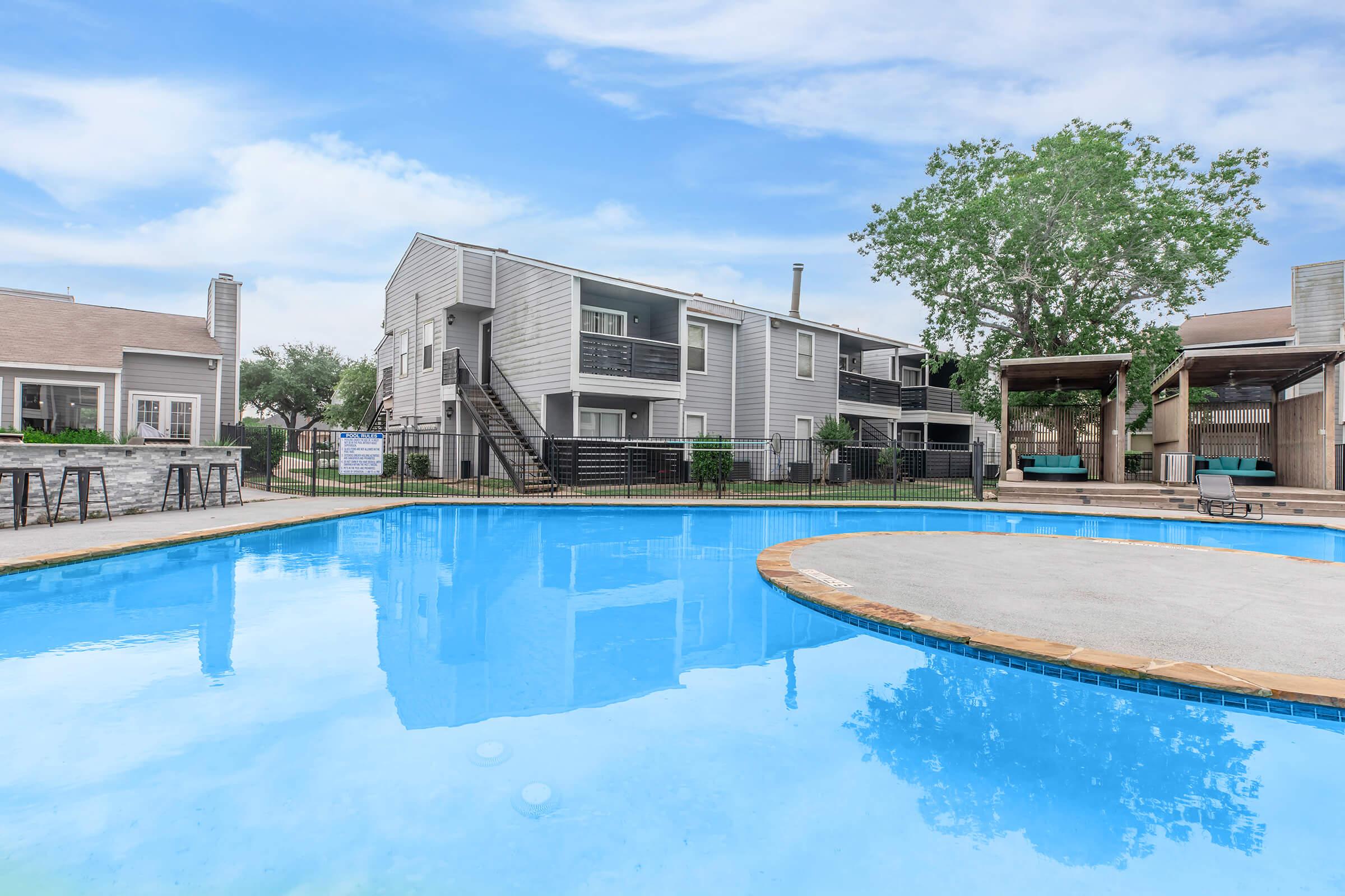 A clear blue swimming pool in an apartment complex with lounge chairs around the edge. In the background, modern two-story buildings and a large tree are visible, along with shaded seating areas beside the pool. The sky is bright with a few clouds, creating a welcoming and relaxing atmosphere.