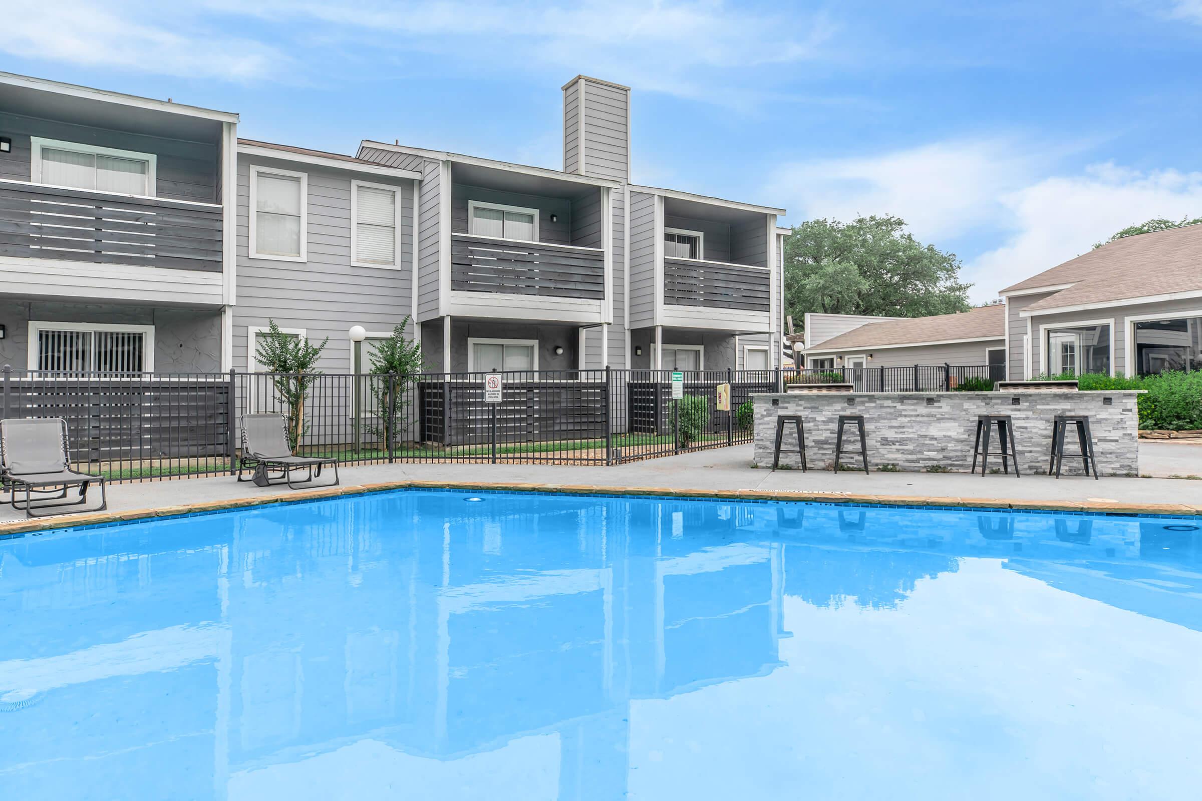 A clear blue swimming pool surrounded by a deck, with several lounge chairs nearby. In the background, there are modern multi-unit buildings with balconies and green trees. A small outdoor bar area is visible next to the pool. The sky is bright blue with a few clouds.