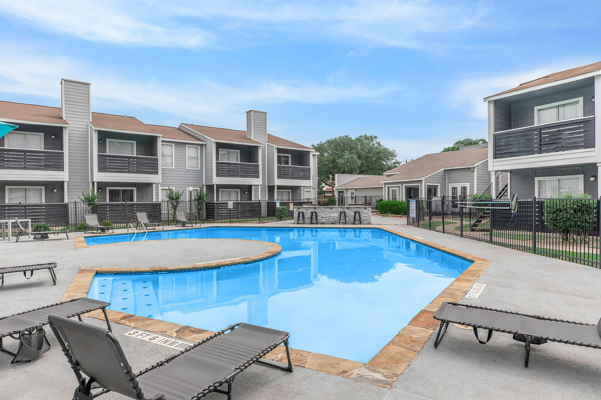 A clear swimming pool surrounded by lounge chairs, with several apartment buildings in the background. The pool area features a shallow section and is enclosed by a black fence. The sky is blue with a few clouds, and there is greenery around the apartments, creating a peaceful residential atmosphere.