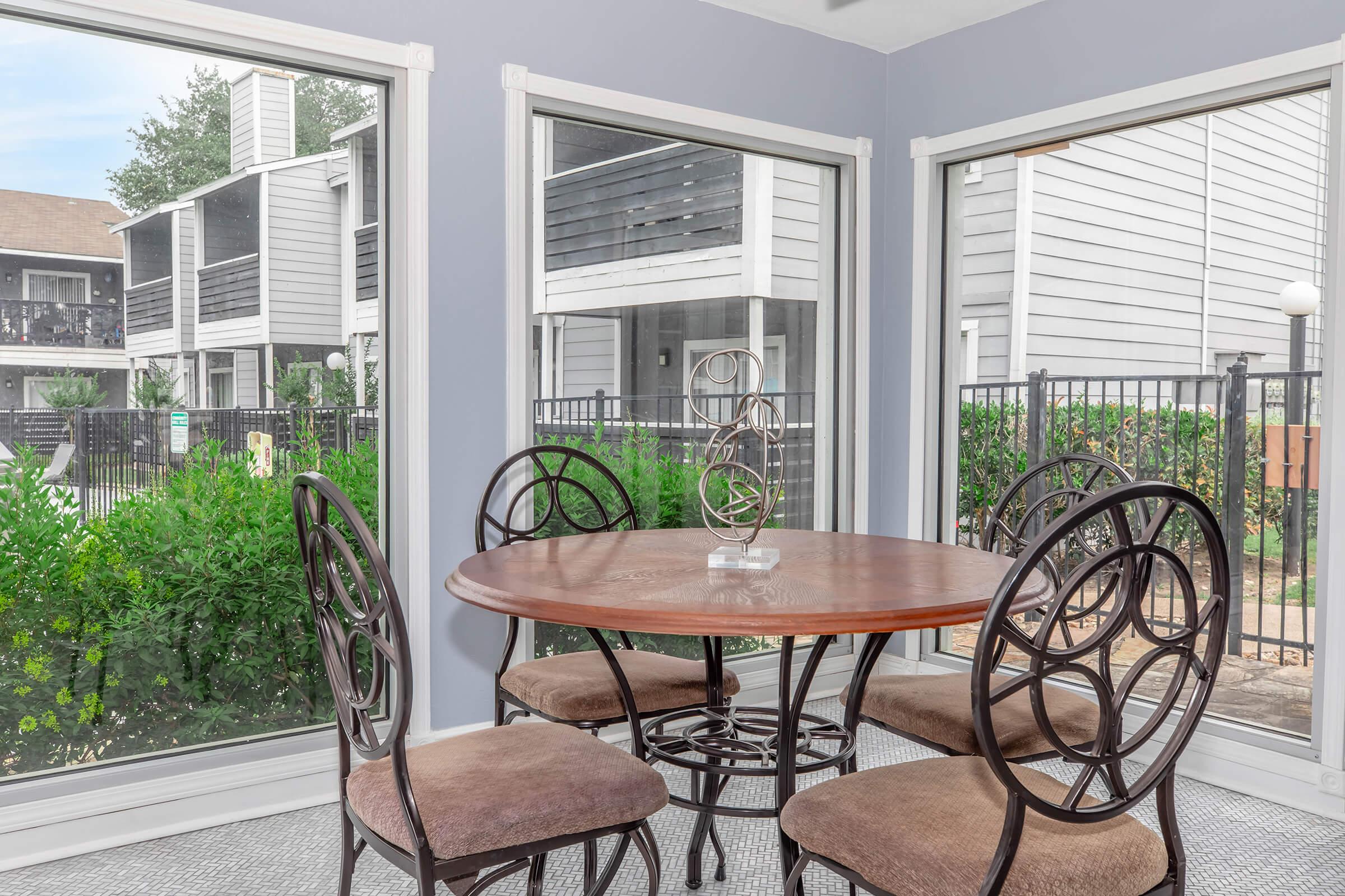 A cozy dining area featuring a round wooden table surrounded by four upholstered chairs, situated in a sunlit space with large windows. Outside, views of lush greenery and an apartment complex can be seen through the glass.
