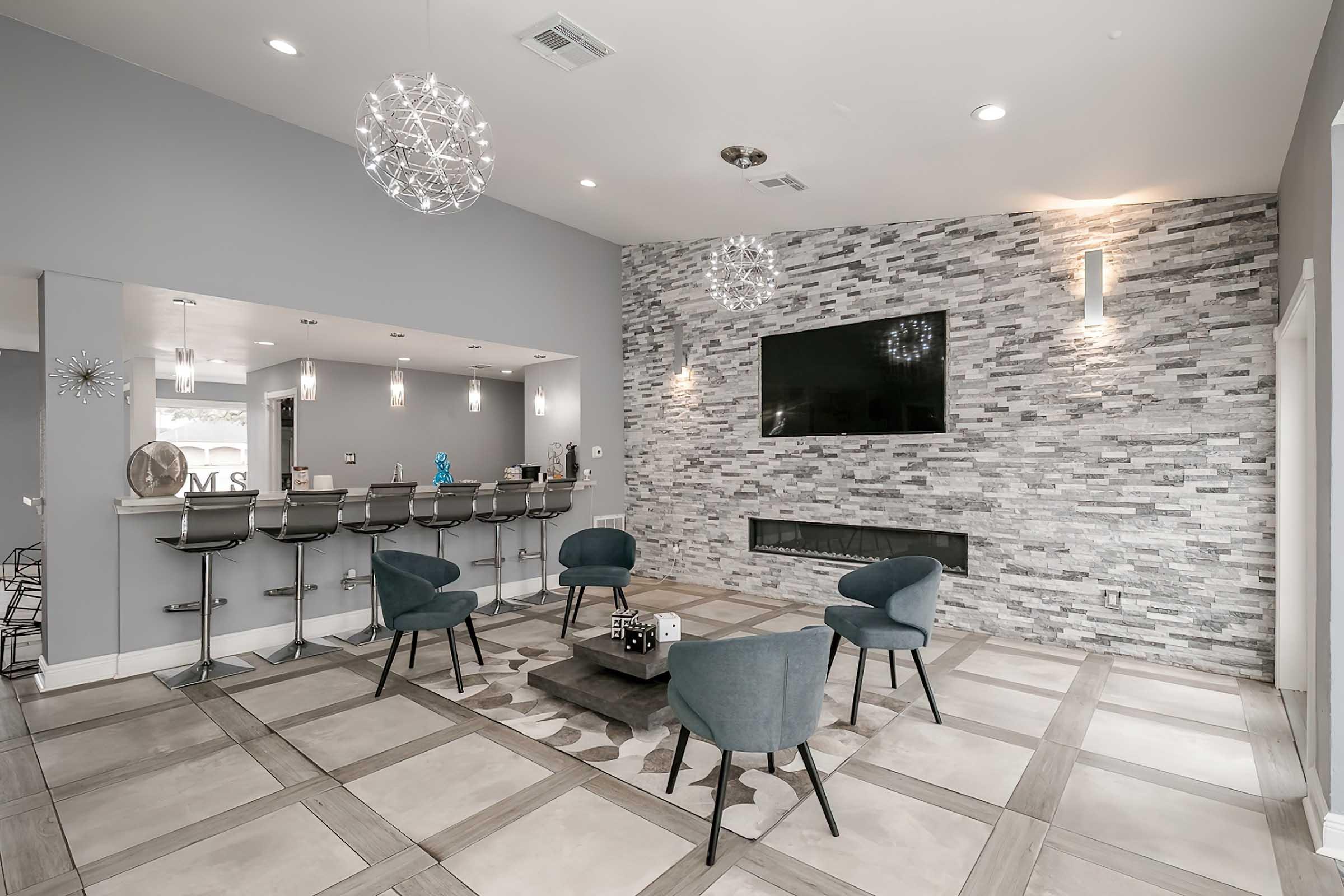 A modern lounge area featuring stylish chairs arranged around a central coffee table, with a bar counter in the background. The space is accentuated by gray walls, decorative lighting, and a stone feature wall with a mounted television. Large windows provide ample natural light, enhancing the inviting atmosphere.
