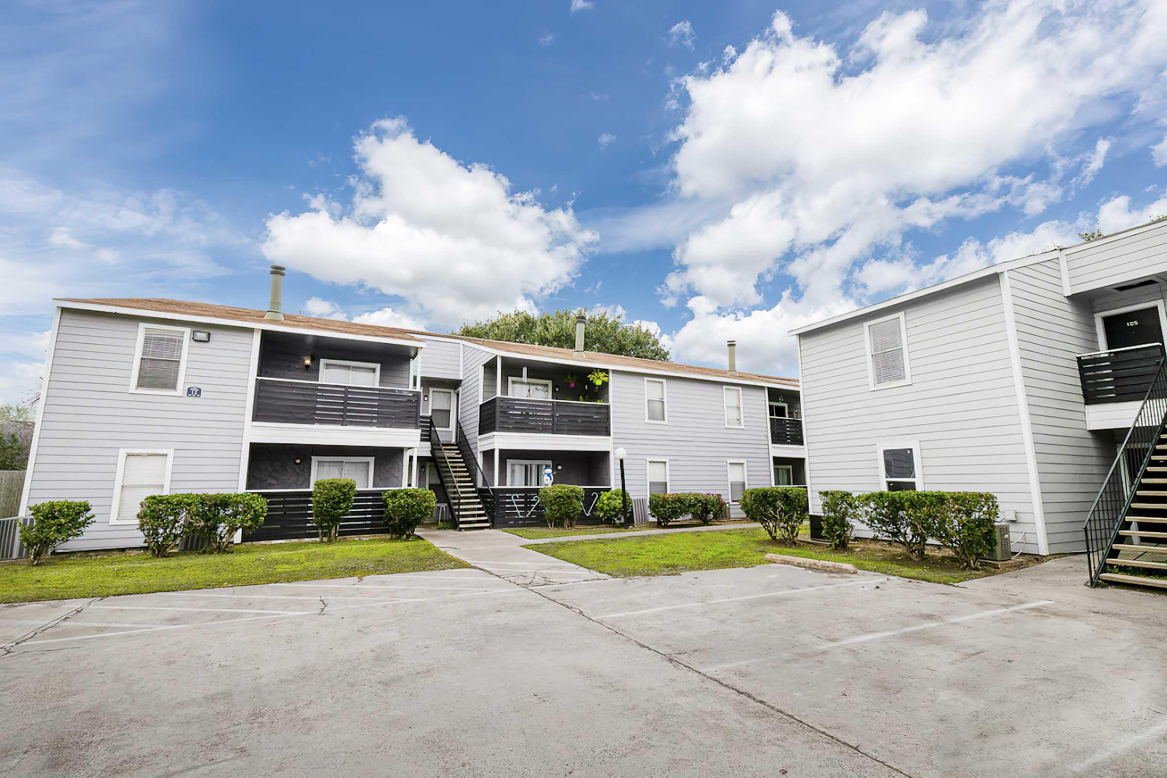 A two-story apartment complex with light gray siding and black staircases. The scene features green lawns and well-maintained shrubs, with a blue sky filled with fluffy white clouds. The pathway leads to the apartment entrances, creating a welcoming residential atmosphere.