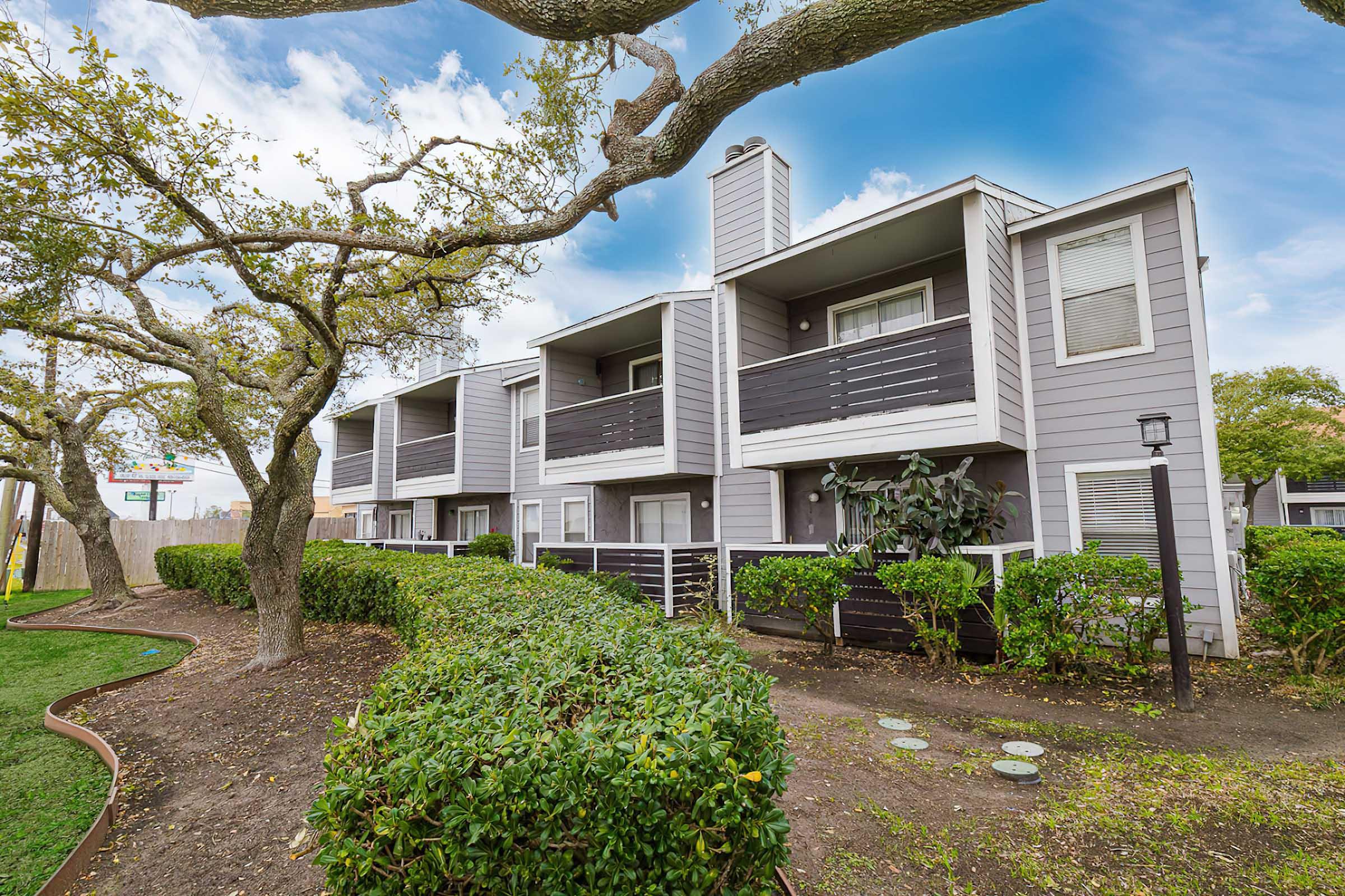 A multi-level apartment building with a gray exterior and black railings, surrounded by well-maintained bushes and trees. The scene includes a pathway and a clear blue sky in the background.