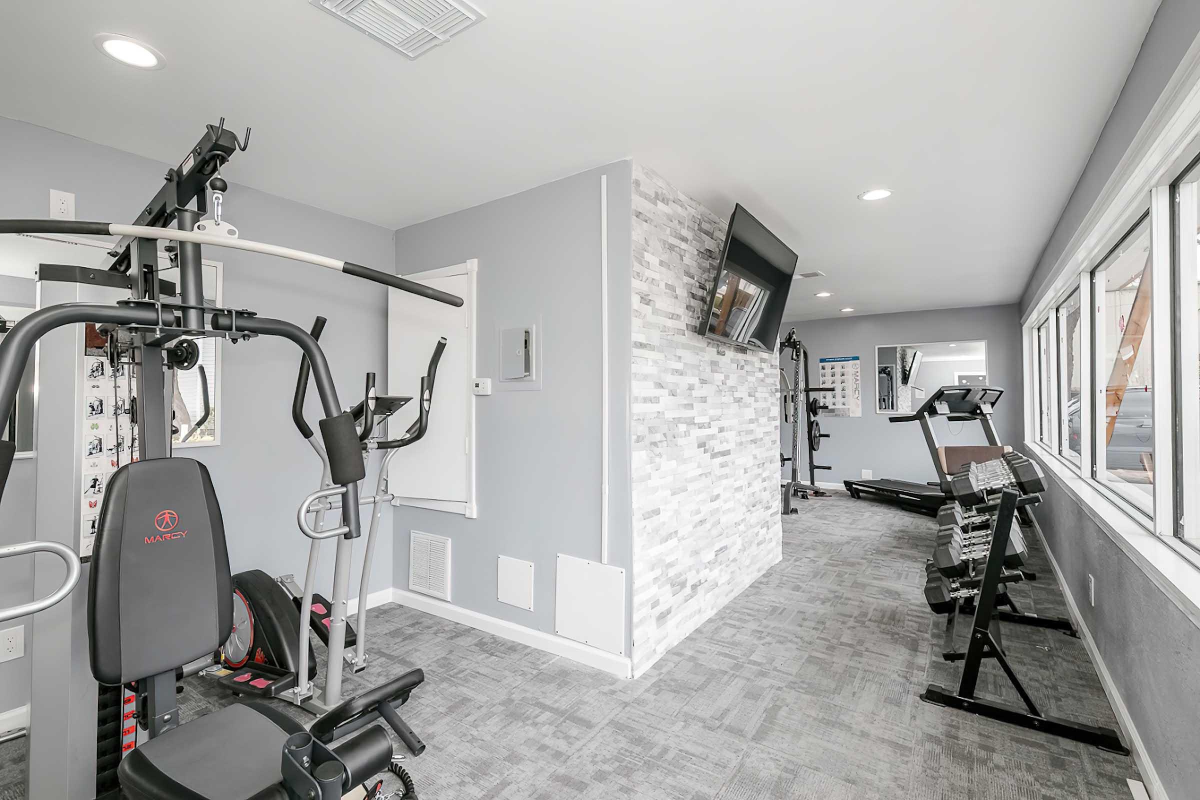 A modern gym interior featuring various exercise equipment, including a multi-station machine, treadmill, and free weights. The space has large windows allowing natural light, with neutral-colored walls and flooring, providing a clean and inviting atmosphere for workouts.