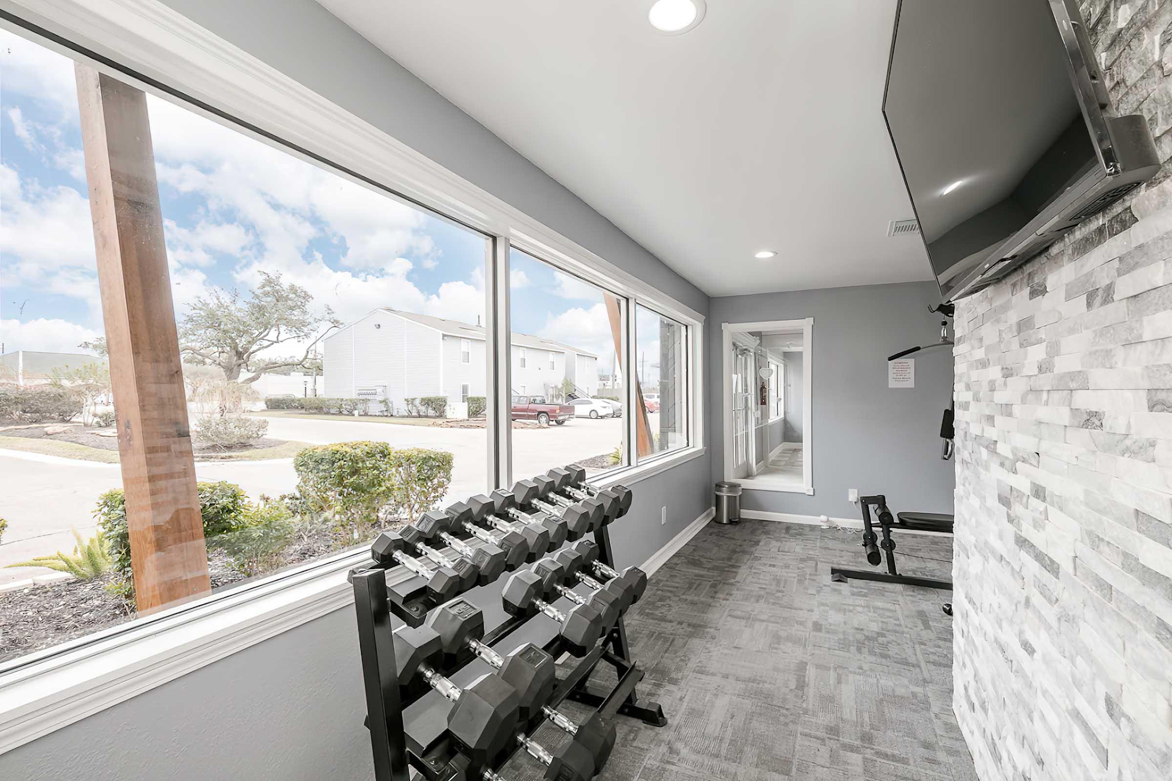 A bright fitness room featuring a rack of dumbbells, a workout bench, and a wall-mounted TV. Large windows provide a view of the outside while letting in natural light, enhancing the workout environment. The decor includes light-colored walls and a textured stone feature wall.