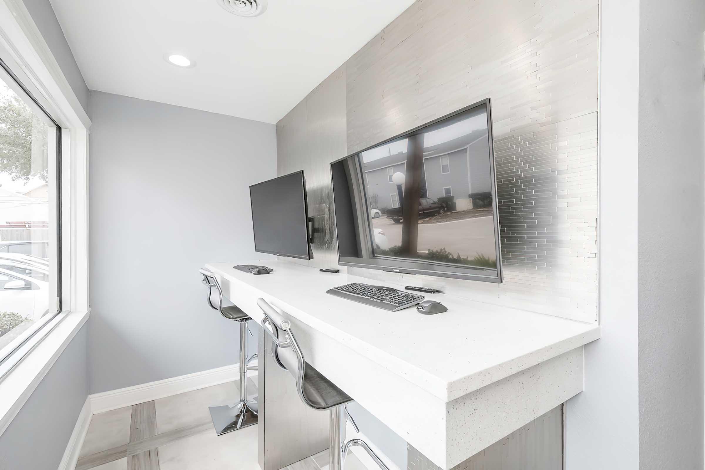 A modern workspace featuring two flat-screen monitors on a sleek white countertop with stools. The walls are a light gray, complemented by a silver panel behind the monitors. Large windows allow natural light to fill the space, creating an open and airy atmosphere.