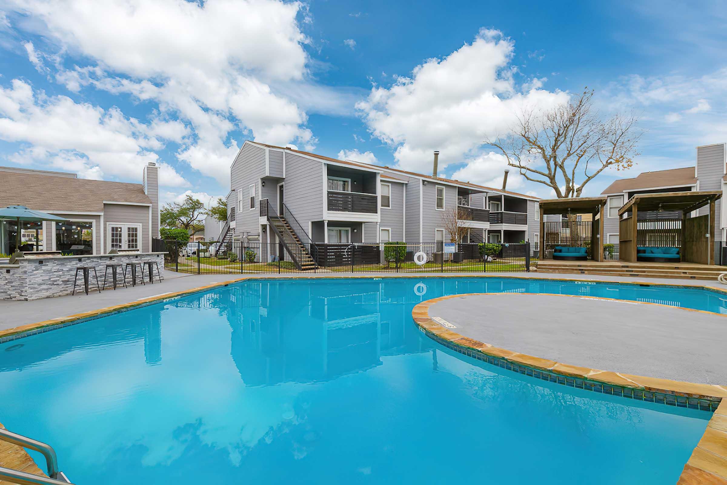 A sparkling blue swimming pool surrounded by landscaped grounds and residential buildings. In the background, apartment units feature balconies, while shaded seating areas are visible near the pool. The sky is bright with fluffy clouds, creating a cheerful atmosphere.