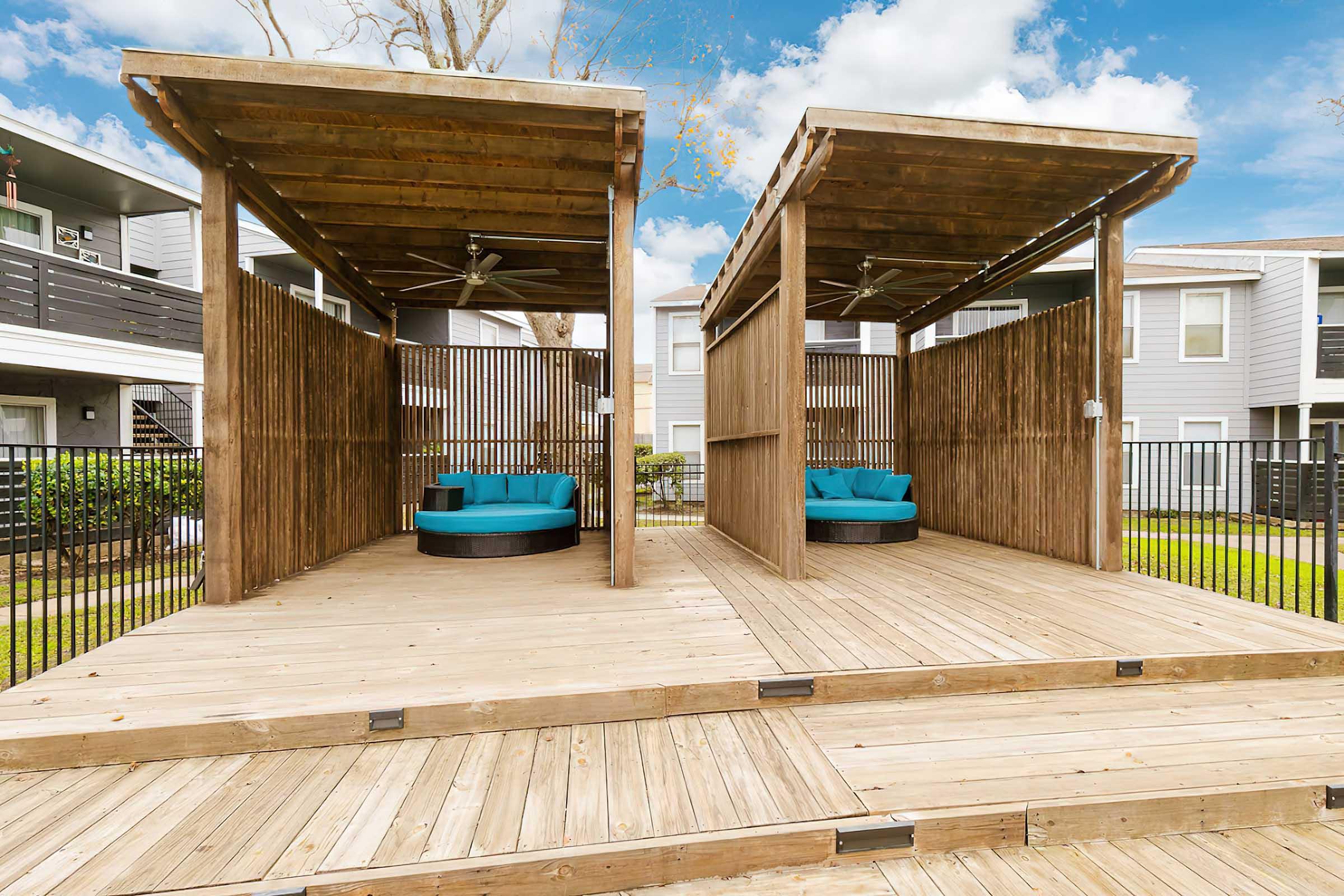 Two wooden cabanas with turquoise seating areas, situated on a deck surrounded by greenery and a fence. The sky is partly cloudy, providing a bright atmosphere. The cabanas are designed for relaxation with fans above for comfort.