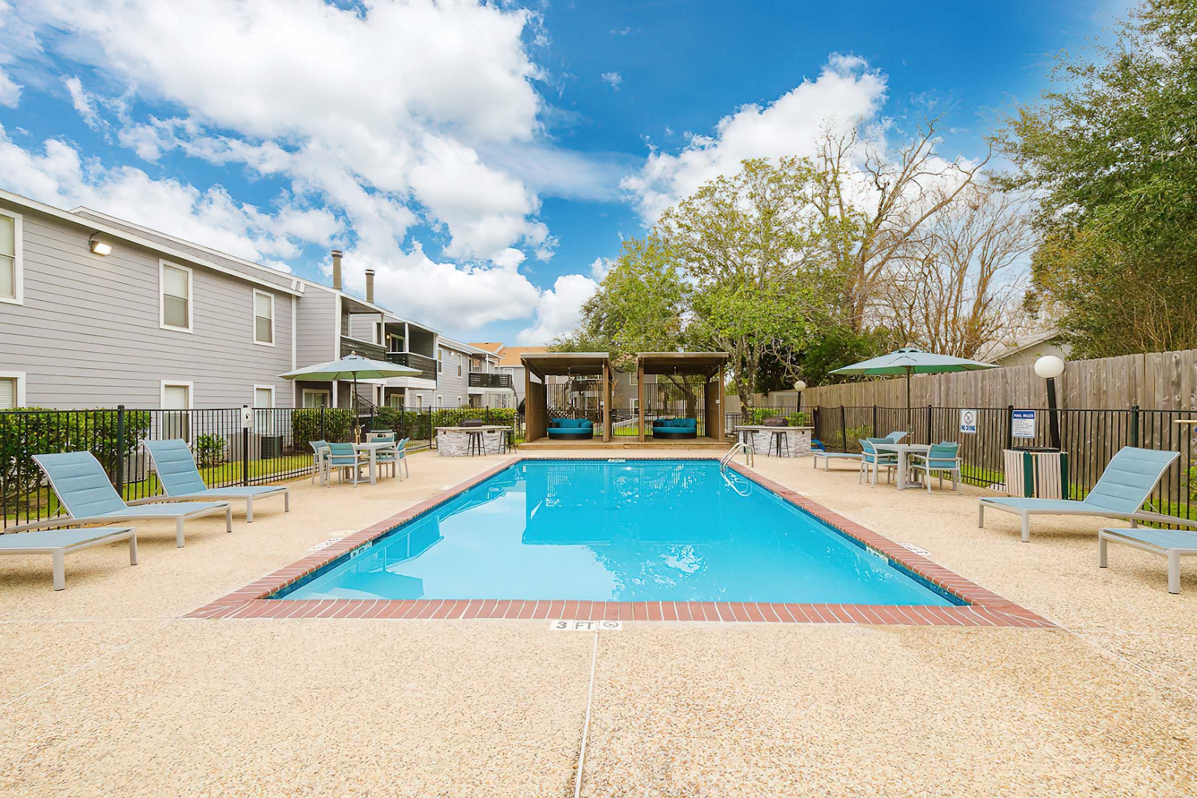Outdoor swimming pool surrounded by lounge chairs and umbrellas. A wooden gazebo is located at one end of the pool area, with a clear blue sky and fluffy white clouds in the background. Nearby buildings are visible, surrounded by greenery and a fenced area.