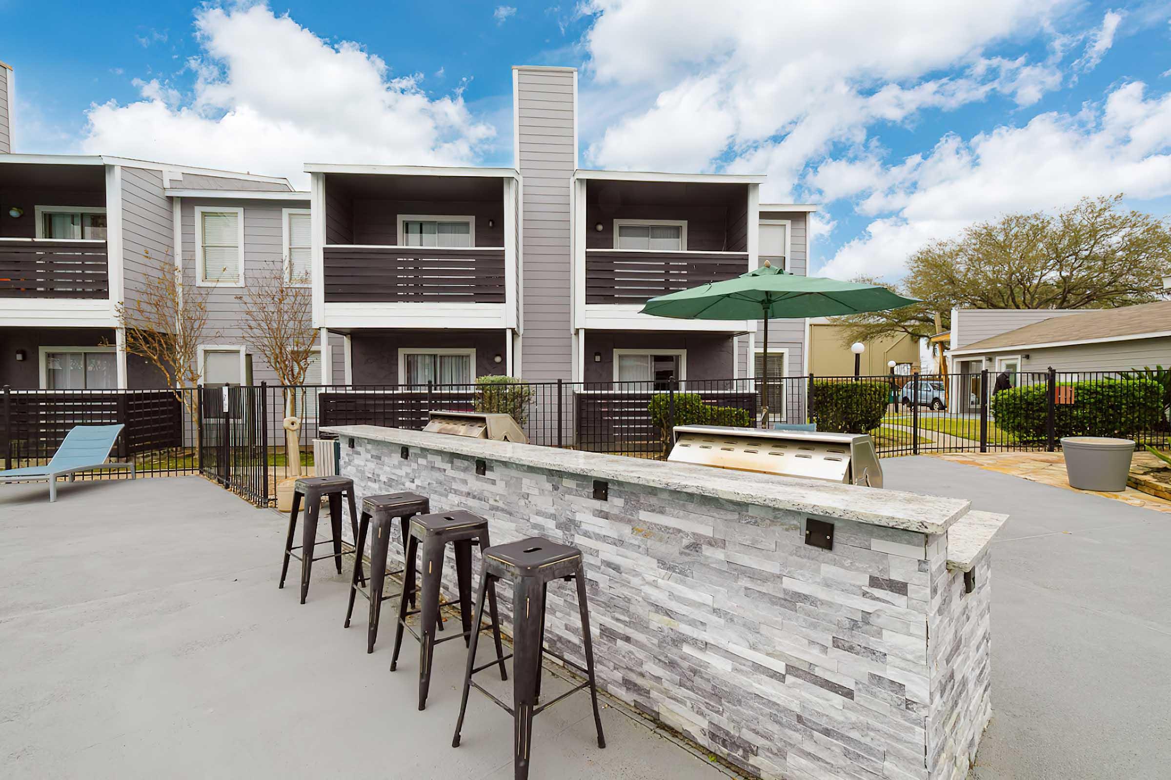 Outdoor area featuring a modern bar with stools, an umbrella, and a grill, surrounded by apartments with a sleek design. Bright blue sky with clouds above, creating a welcoming atmosphere. The space is fenced, enhancing privacy and comfort for gatherings.