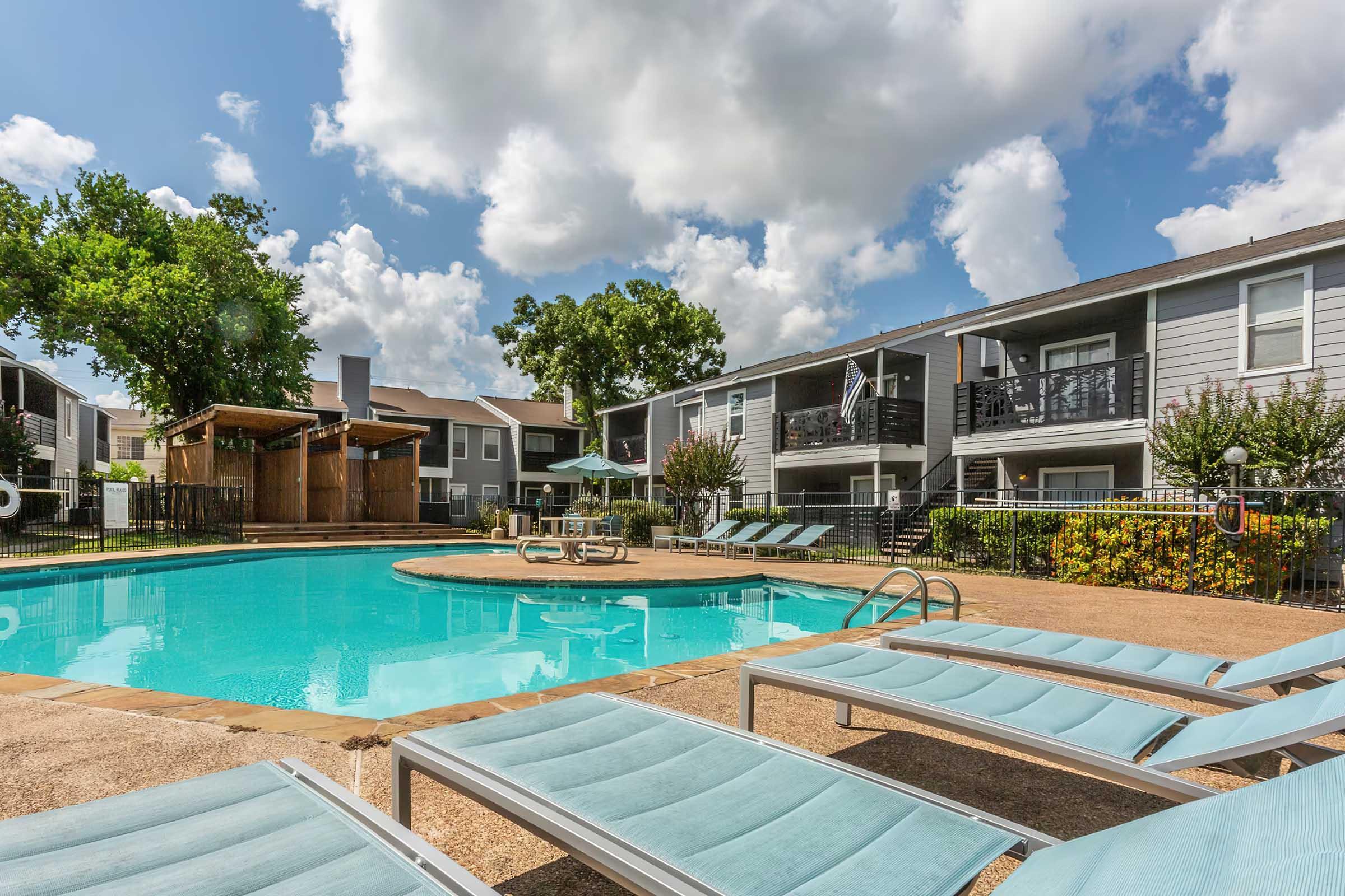 A tranquil swimming pool surrounded by lounge chairs, with apartment buildings in the background. Lush greenery and sunny skies enhance the relaxing atmosphere, ideal for leisure and outdoor enjoyment.