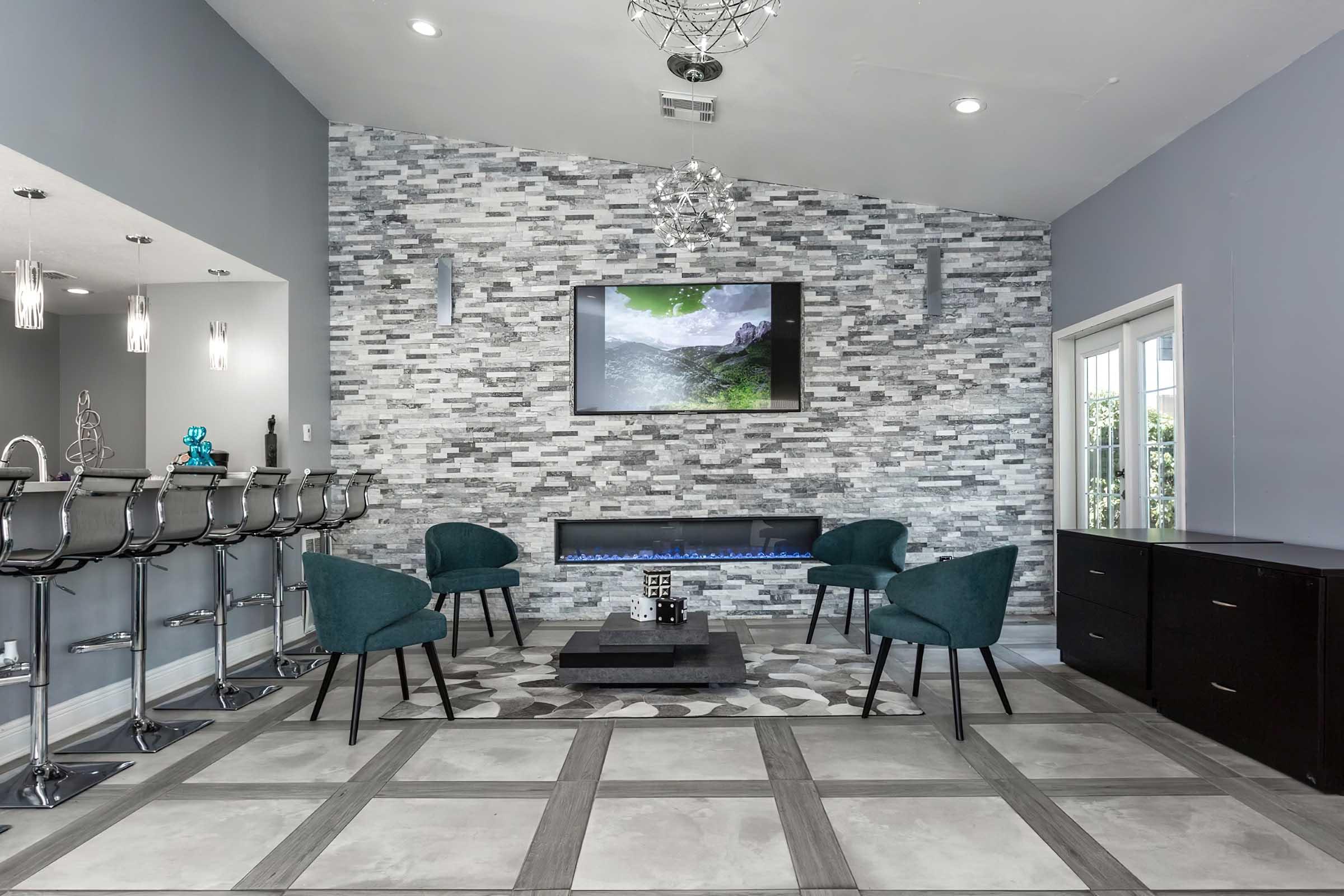 A modern living space featuring a stone accent wall with a mounted television, a sleek electric fireplace, and four green chairs arranged around a dark coffee table. A bar area with stylish stools is visible on the left, and large windows illuminate the room, enhancing its contemporary design.