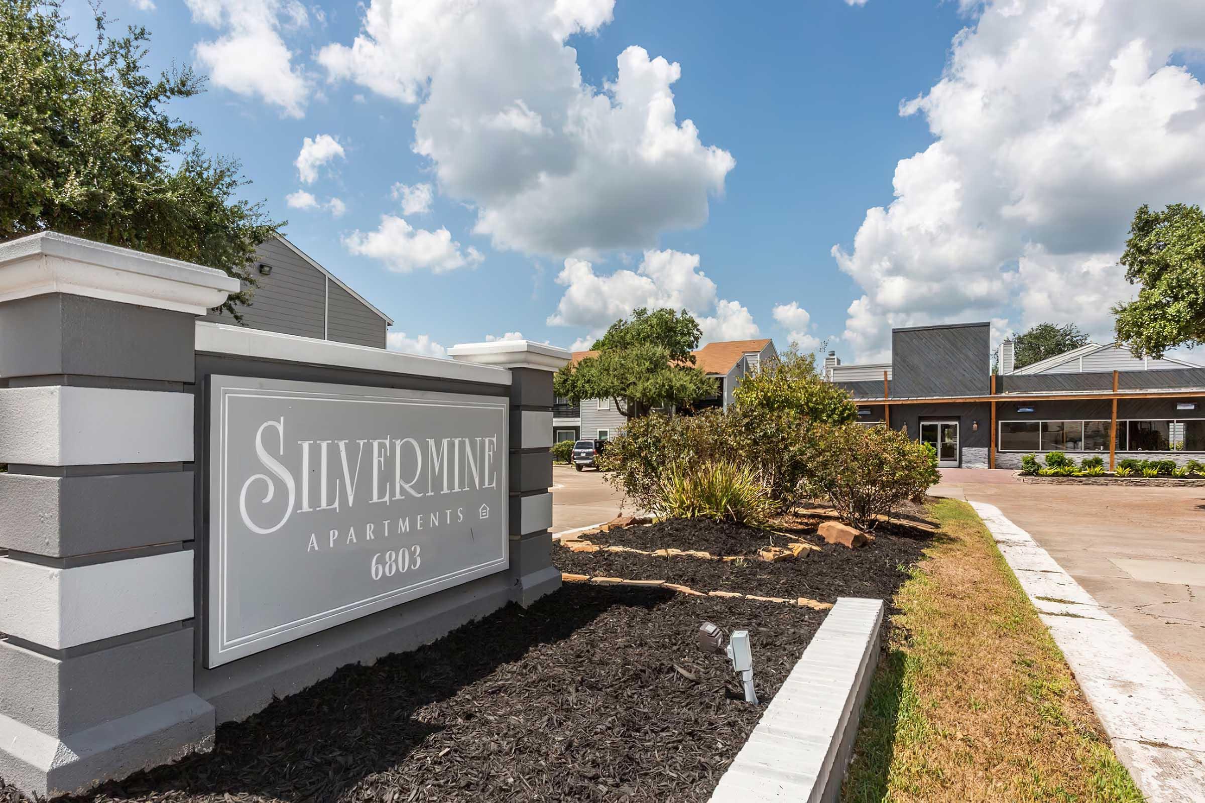 Sign for Silvermine Apartments located at 6803, featuring a landscaped entrance with shrubs and mulch. The background includes buildings and a bright blue sky with fluffy clouds.
