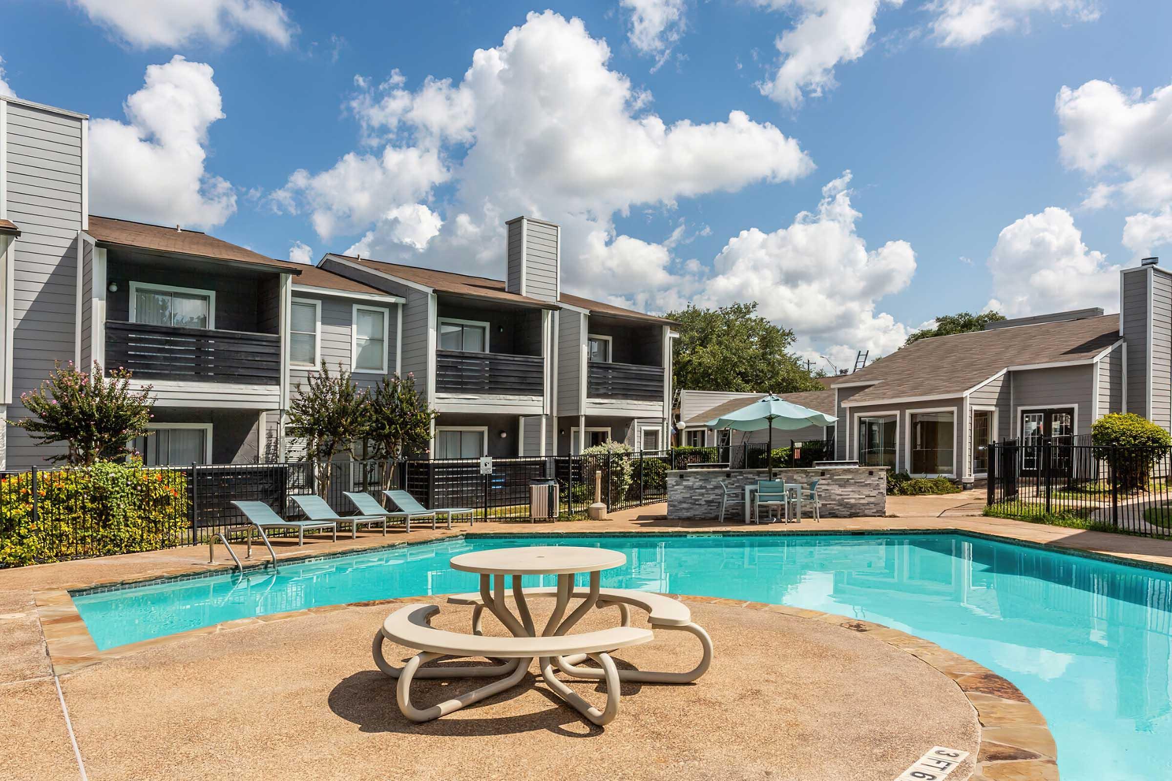 A bright, sunny day at a residential complex featuring a clear blue swimming pool surrounded by lounge chairs and landscaped greenery. In the background, several multi-story buildings with balconies are visible, under a partly cloudy sky. A shaded seating area is positioned near the pool.