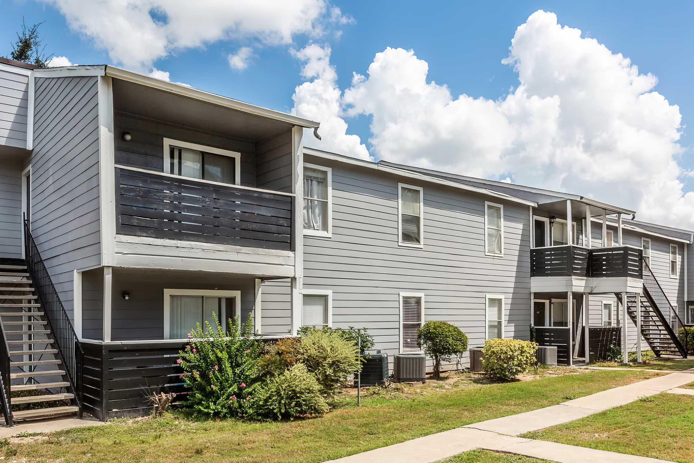 A modern apartment complex featuring two-story buildings with gray siding. Each unit has a balcony, and there are staircases leading to the upper levels. The landscape includes neatly trimmed bushes and patches of grass, set against a bright blue sky with fluffy white clouds.