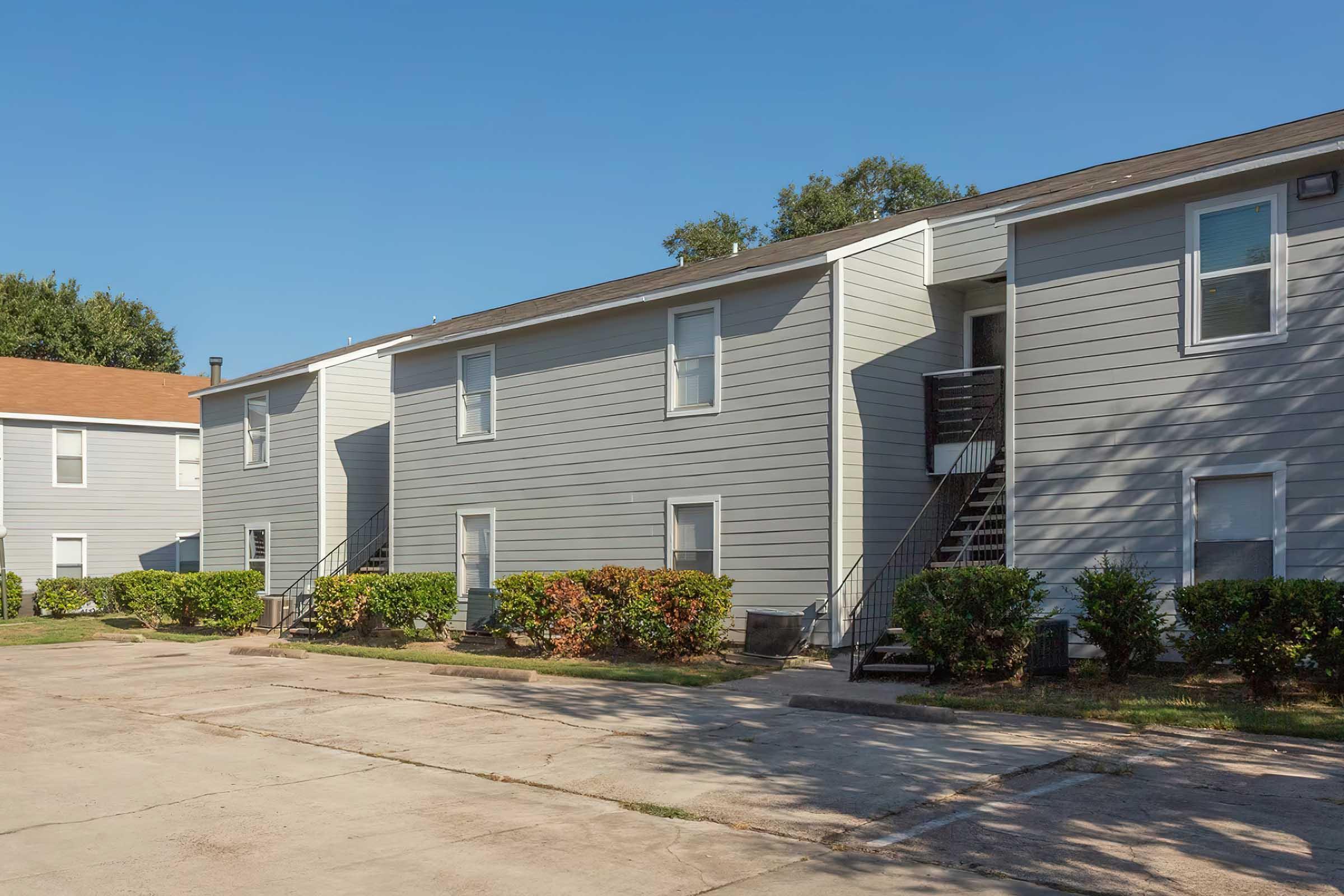 Two gray apartment buildings with a paved parking area in front. The buildings have multiple units, each with stairs leading to the second floor. Well-maintained shrubs line the walkways. The sky is clear and blue, suggesting a sunny day.