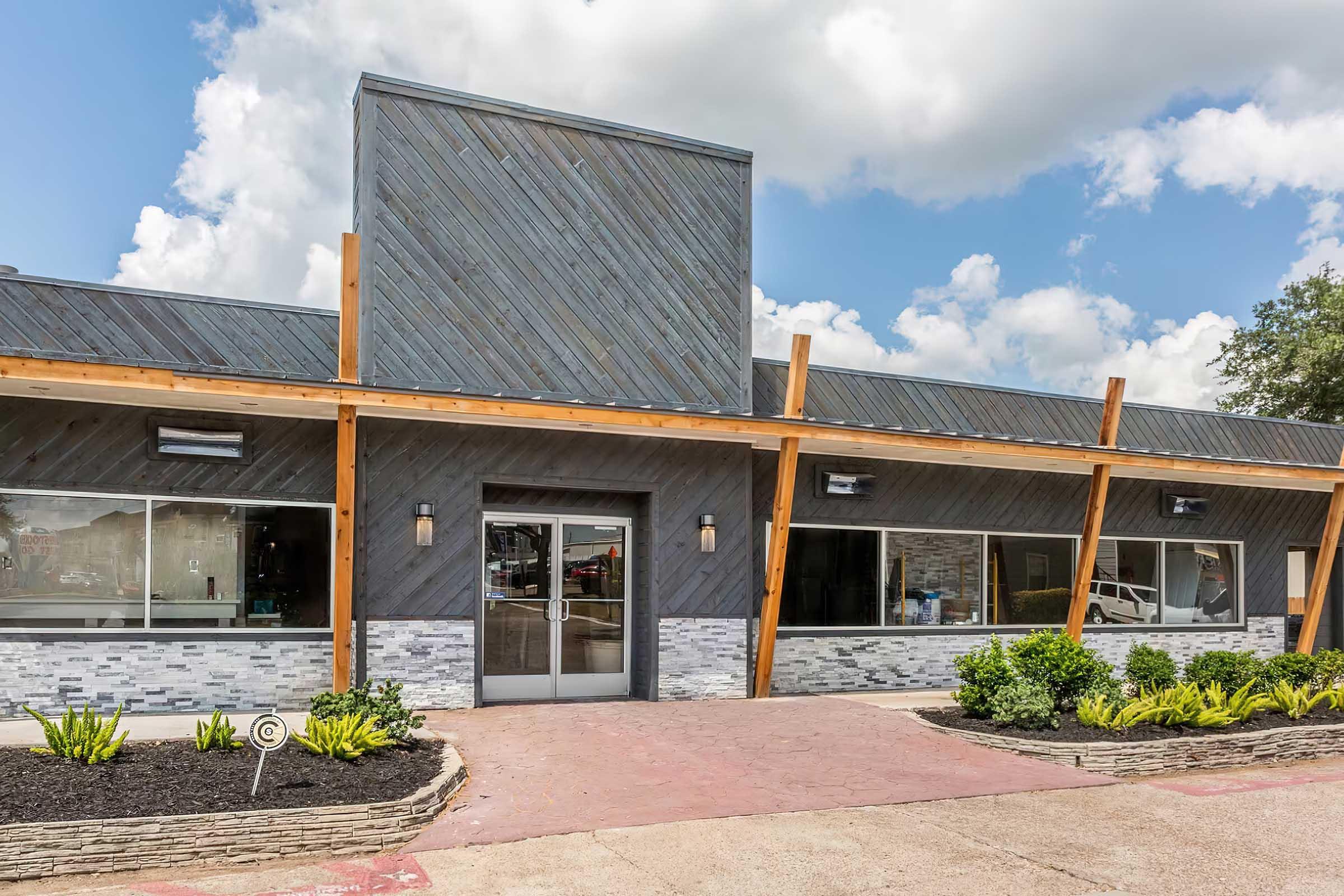 A modern building with a sleek design featuring a combination of wood and stone accents. The structure has large windows, a glass entrance door, and decorative landscaping with shrubs. The sky is partly cloudy, adding a bright atmosphere to the scene.