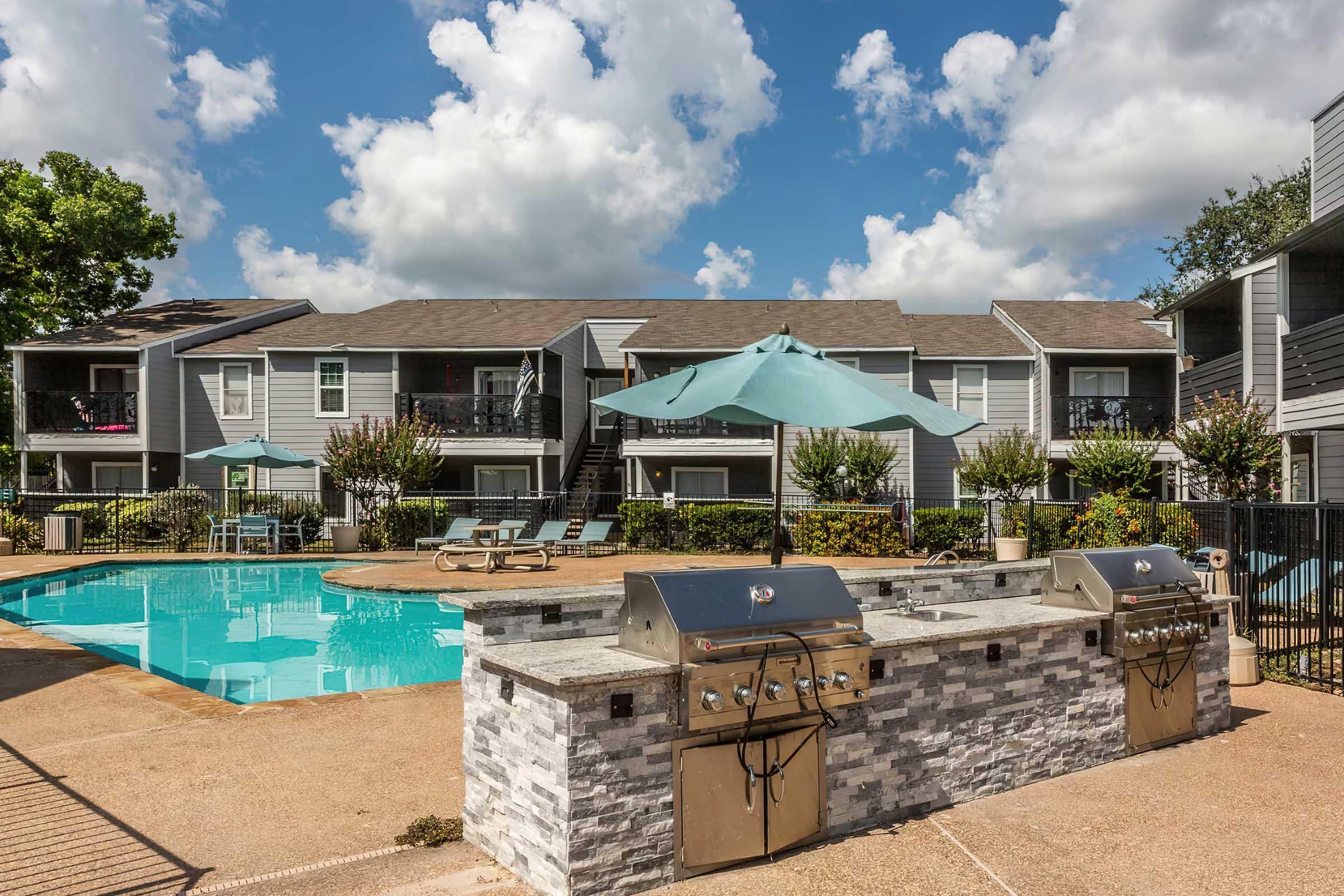 A well-maintained apartment complex featuring a pool surrounded by lounge chairs, with outdoor grills and an umbrella for shade. Lush greenery and flowers add to the pleasant atmosphere under a bright blue sky with fluffy clouds.