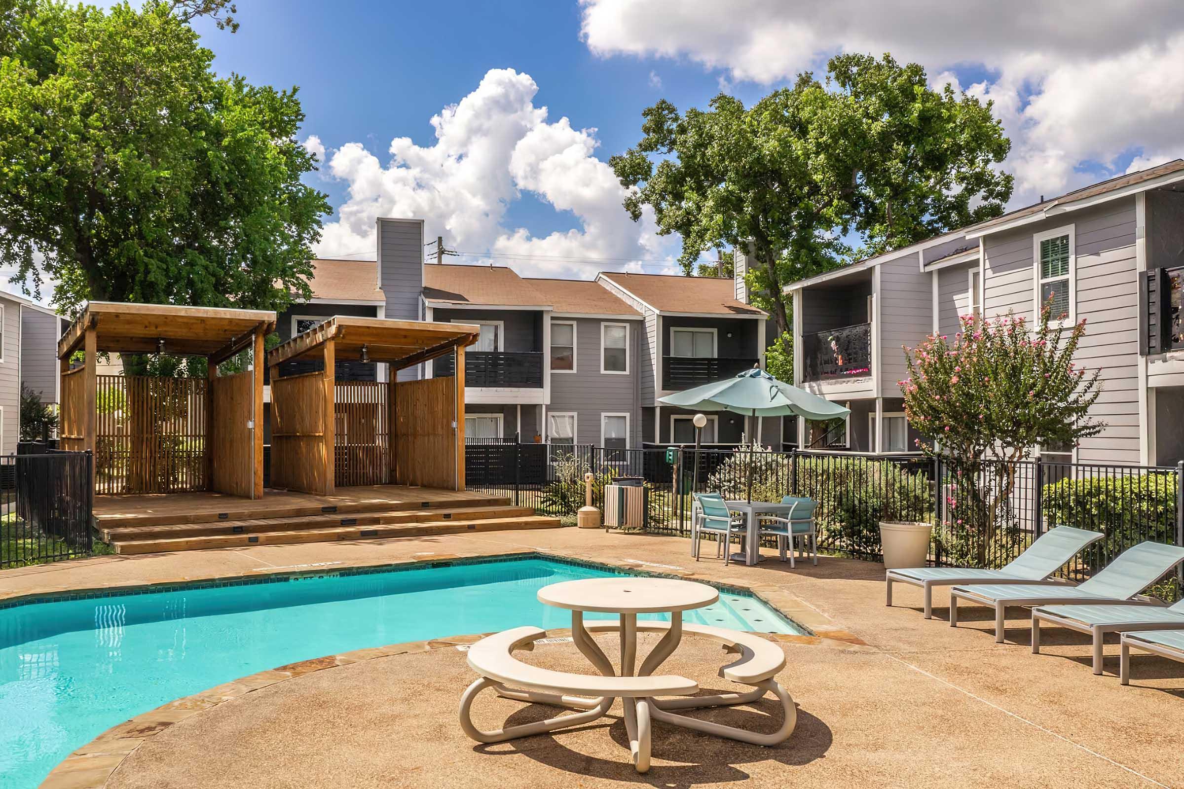 A residential pool area featuring a blue swimming pool with lounge chairs around it, a wooden pergola, and nearby patio furniture. The background includes two apartment buildings surrounded by trees under a partly cloudy sky.