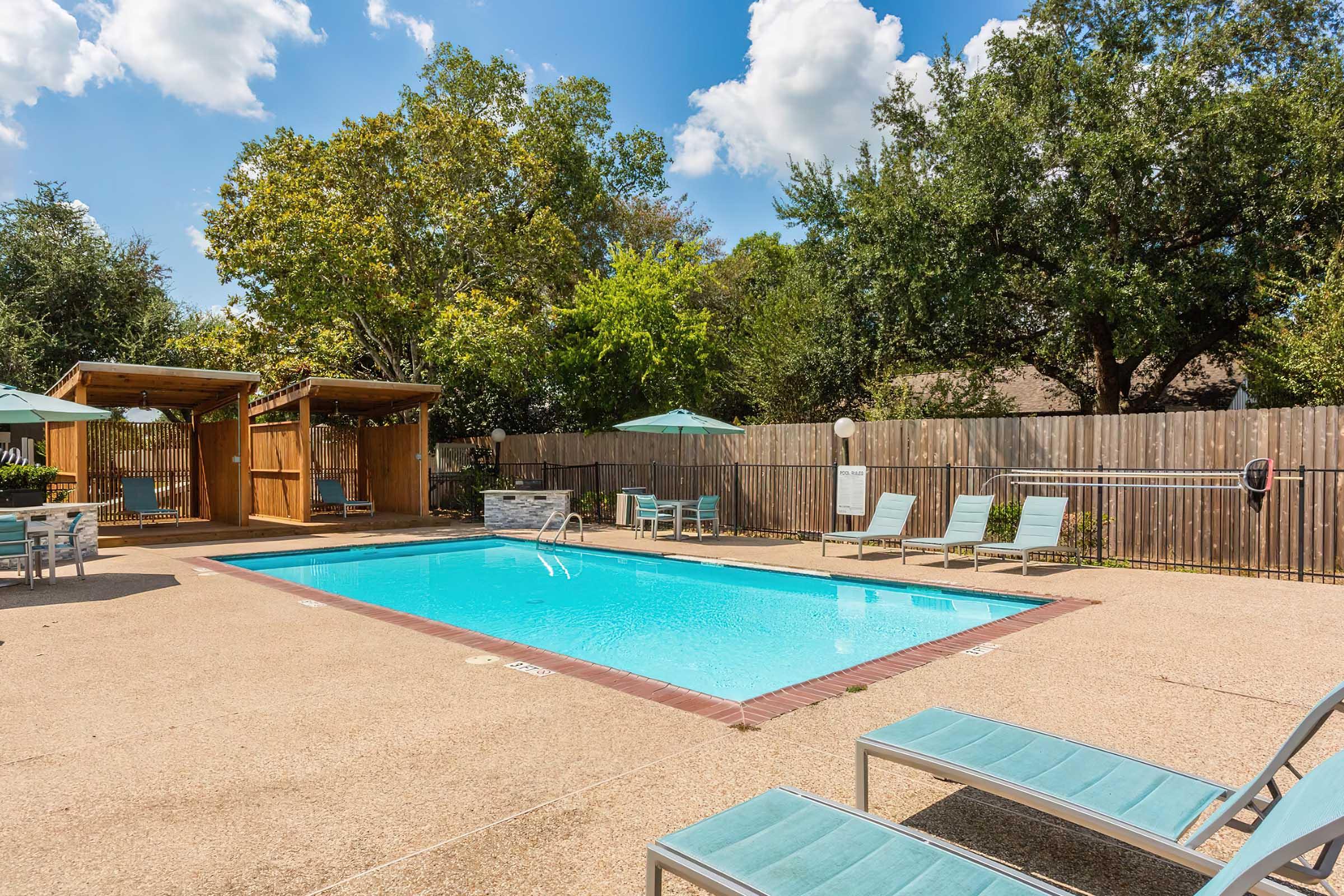 A sunny outdoor pool area with clear blue water. Several lounge chairs are arranged around the pool, and two shaded cabanas are visible. Lush green trees and a wooden fence surround the area, creating a relaxing atmosphere. Bright blue sky with a few fluffy clouds overhead.