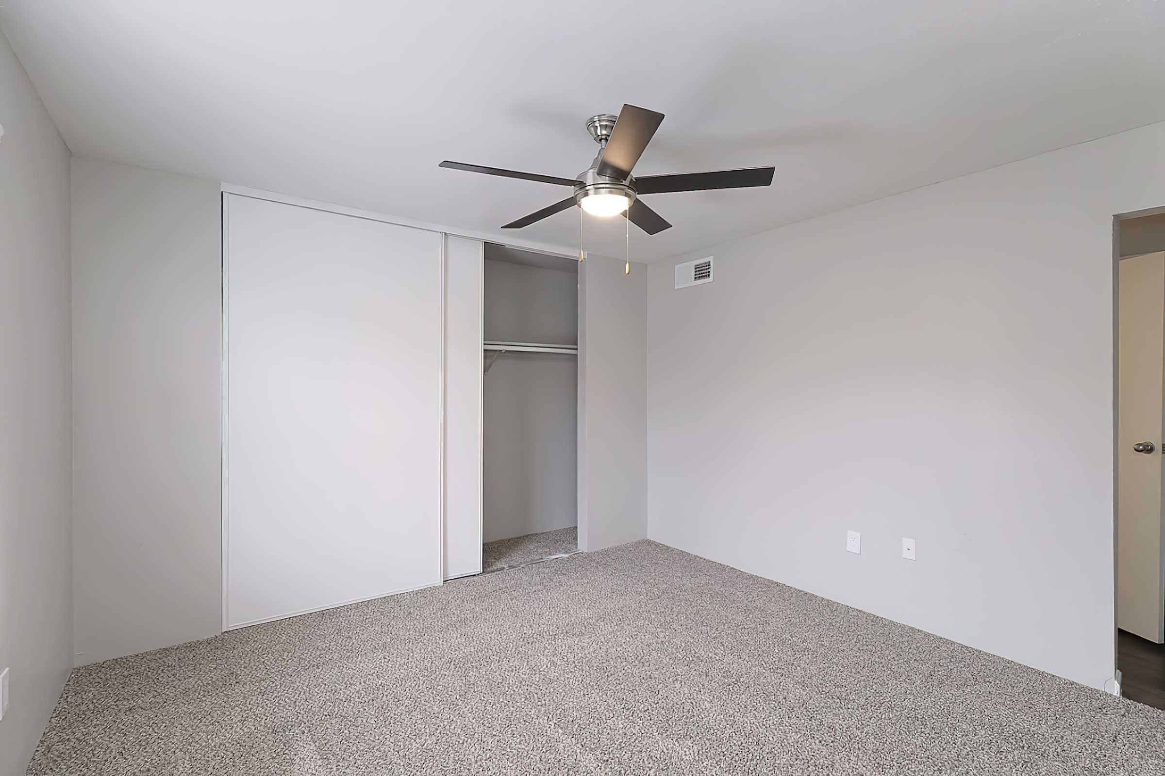 A spacious room featuring light gray walls and a ceiling fan. The floor is carpeted, and there is a white sliding closet door on one side. The room is well-lit, creating a bright atmosphere, and there is an additional entrance to another space on the right.