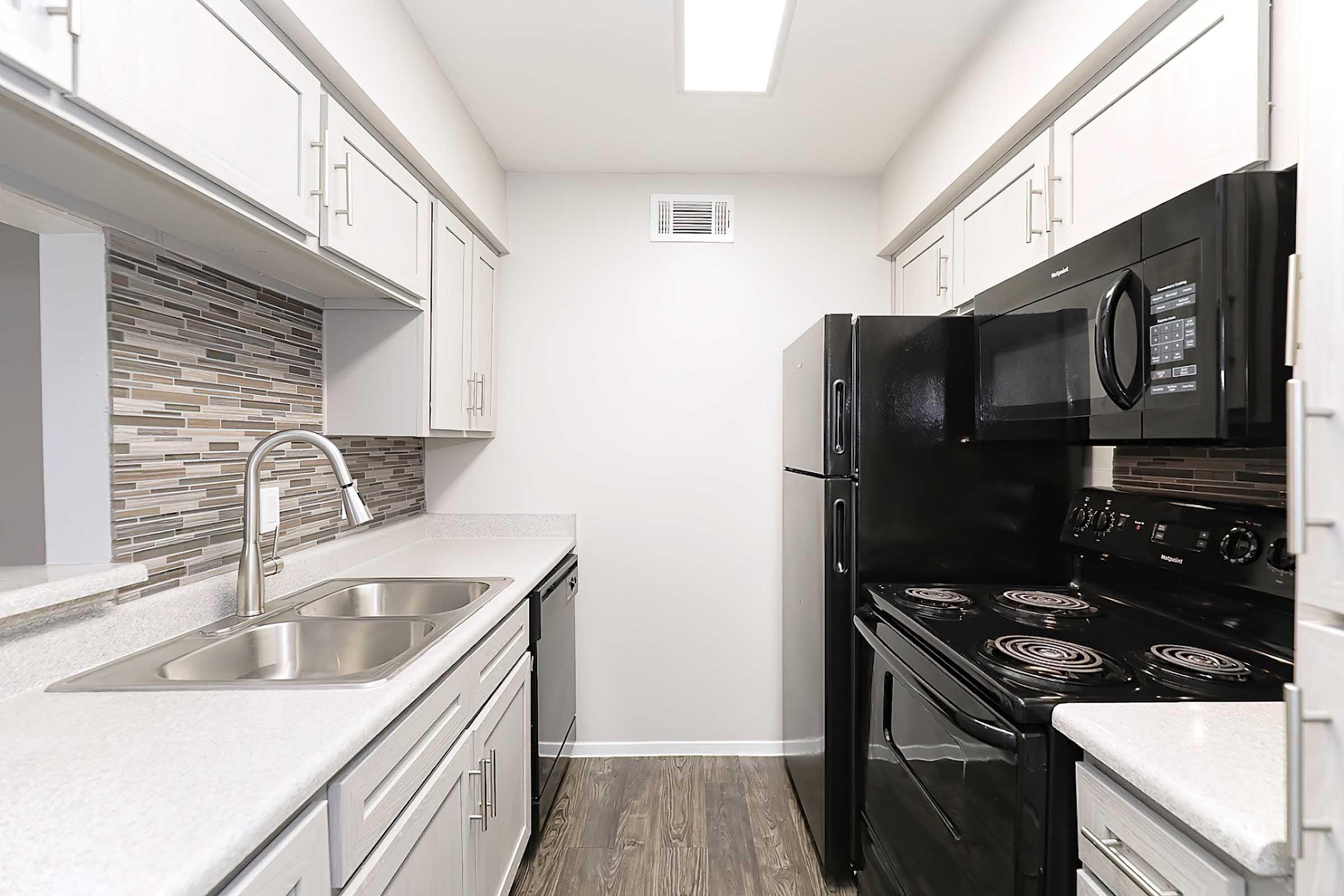 Modern kitchen featuring white cabinets, a double sink, and black appliances including a refrigerator and an oven. The backsplash has a mosaic pattern, and the space is well-lit with a ceiling fixture. The flooring is a dark wood laminate, creating a stylish contrast.