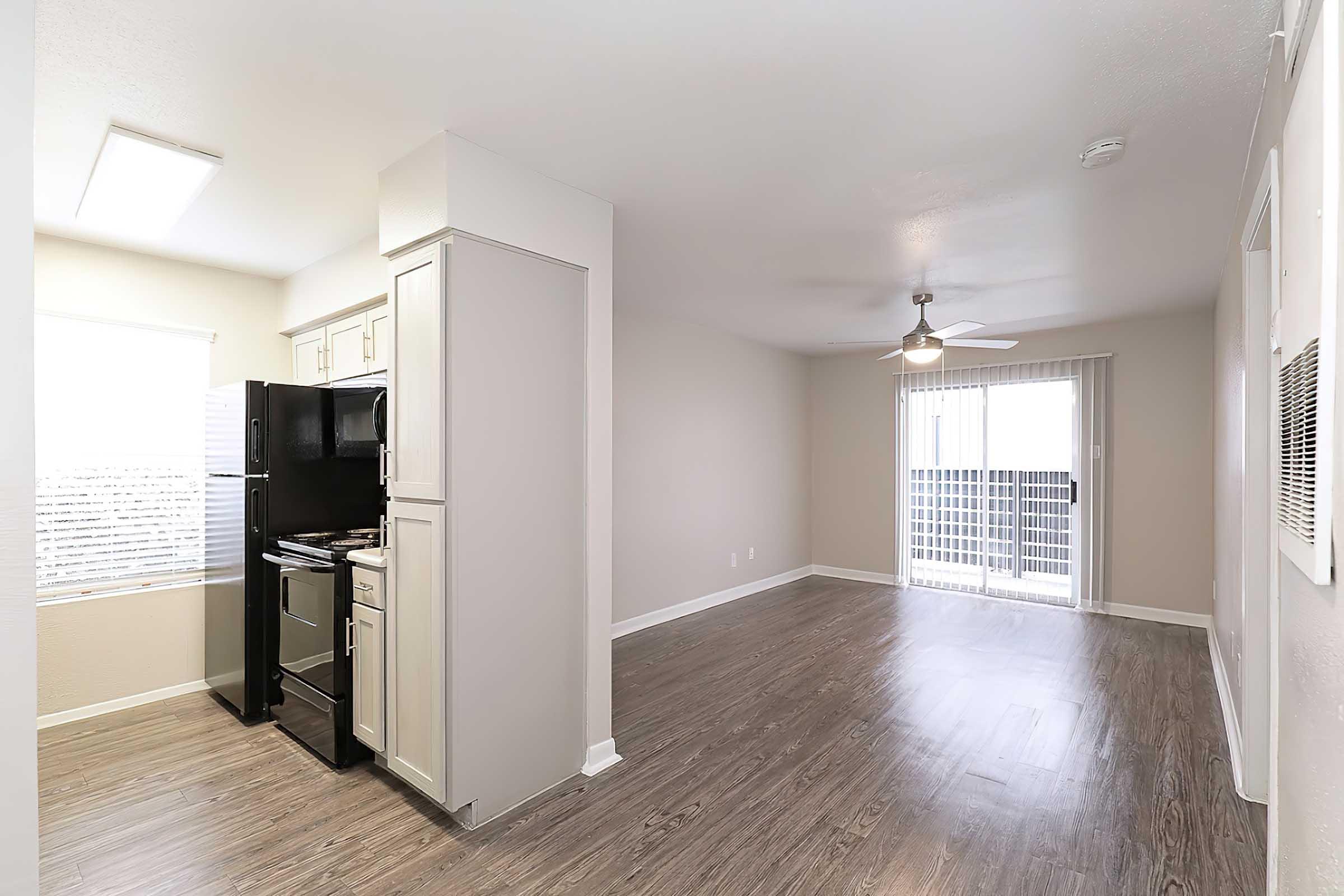 Modern open-concept kitchen and living area featuring light-colored walls, a ceiling fan, and large windows. The kitchen includes black appliances and cabinets, while the living space has wood-style flooring and access to a balcony through sliding glass doors.
