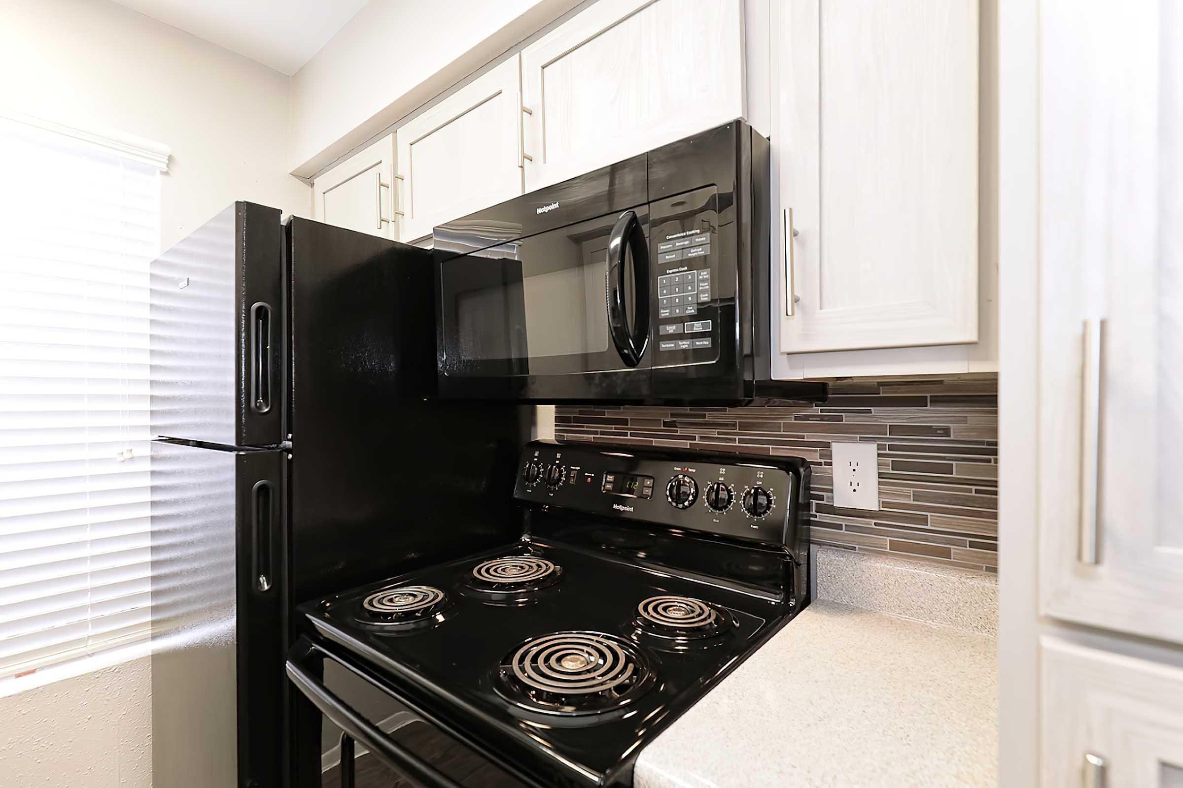 A modern kitchen featuring a black refrigerator and a black stove with an oven, alongside a built-in microwave. The backsplash consists of gray tiles, and there are white cabinets above the countertop. Natural light filters through a window with blinds.