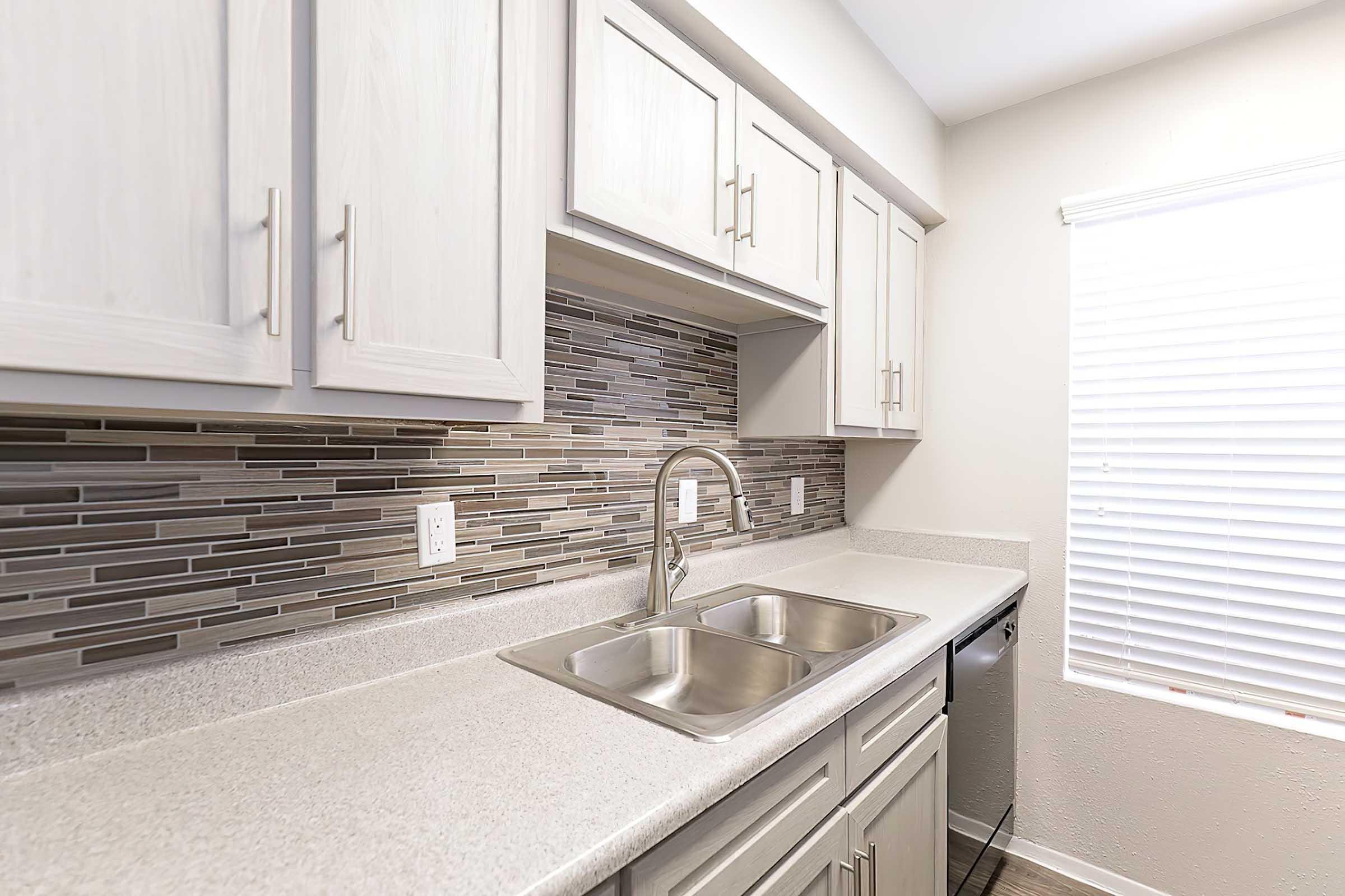 Light-colored kitchen featuring modern cabinets and a dual sink. The backsplash is composed of stylish gray and beige tiles, while natural light enters through a window with white blinds. A sleek counter space is visible, enhancing the contemporary feel of the kitchen.