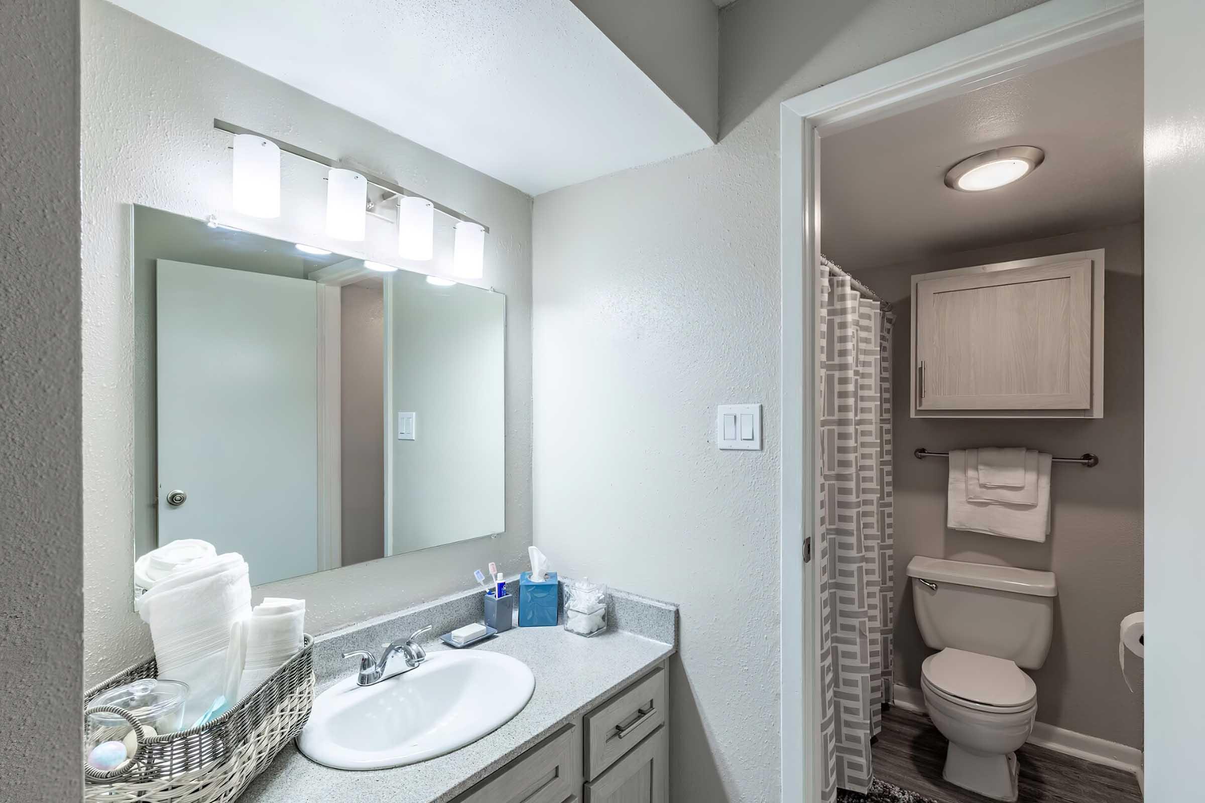 A clean and modern bathroom featuring a white sink with a mirror above it, a shower with a patterned curtain, a toilet, and light-colored cabinets. The countertop is adorned with neatly arranged toiletries and a basket of towels. Soft lighting enhances the inviting atmosphere.