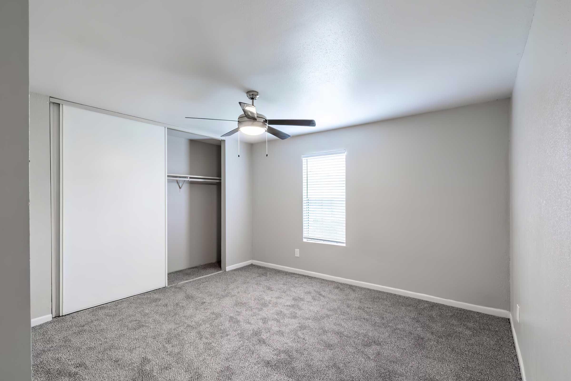 A spacious, empty bedroom featuring light gray walls and a plush carpet. The room includes a ceiling fan, a window with blinds, and a closet with sliding doors. Soft natural light illuminates the space, making it feel bright and inviting.