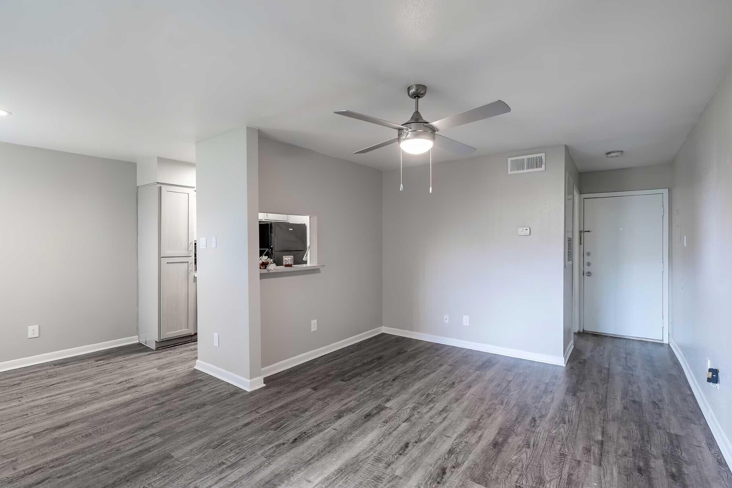An empty room with light gray walls and a ceiling fan. The floor has laminate wood-like finish. There’s a doorway leading to the entrance on the right and an open space that connects to a kitchen area with light-colored cabinetry in the background. Natural light fills the space.