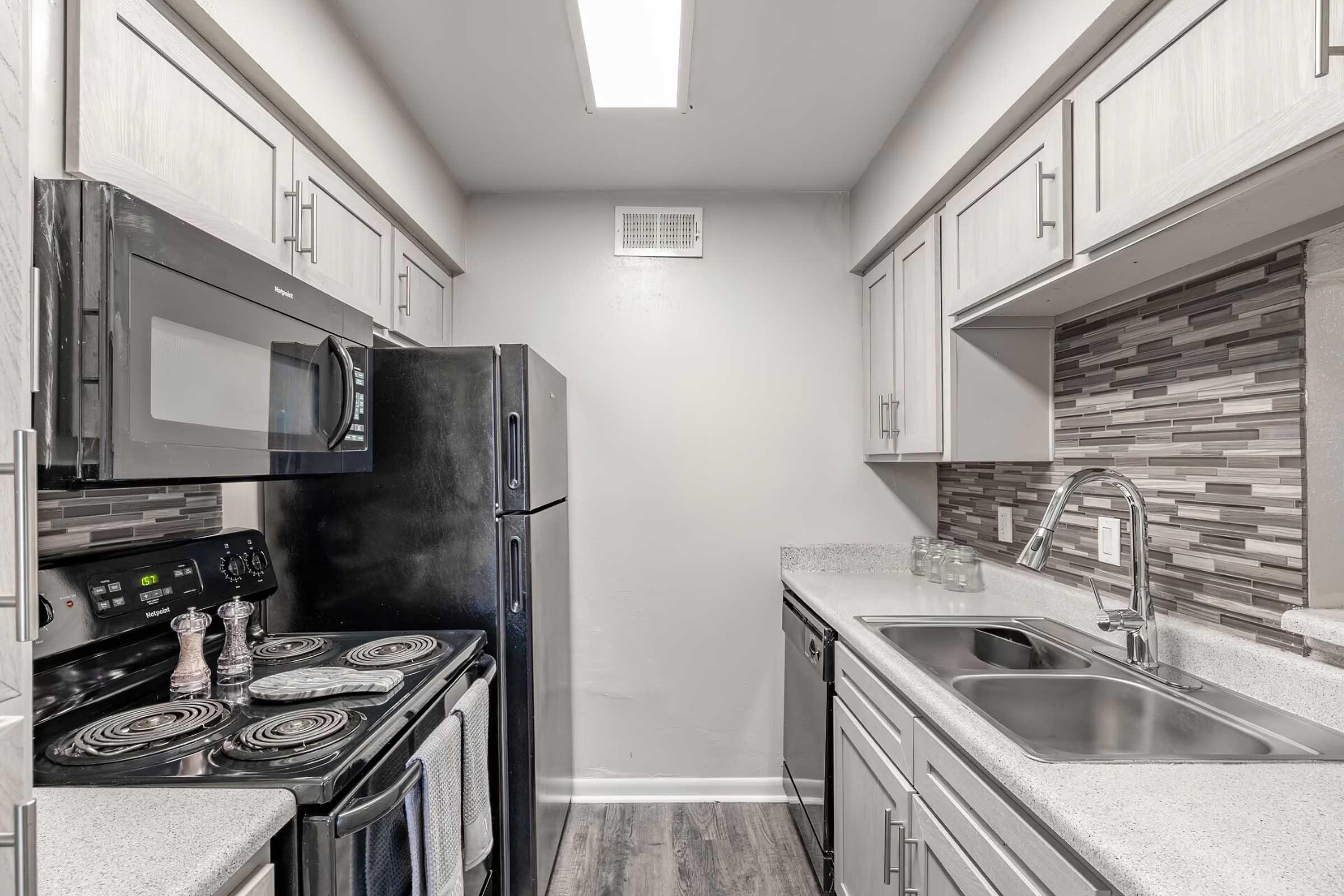 A modern kitchen featuring light gray cabinets, a black refrigerator, and a black stove with an overhead microwave. The countertops are speckled gray with a tiled backsplash in shades of gray. The kitchen is well-lit with a ceiling light and has a double sink next to the dishwasher.