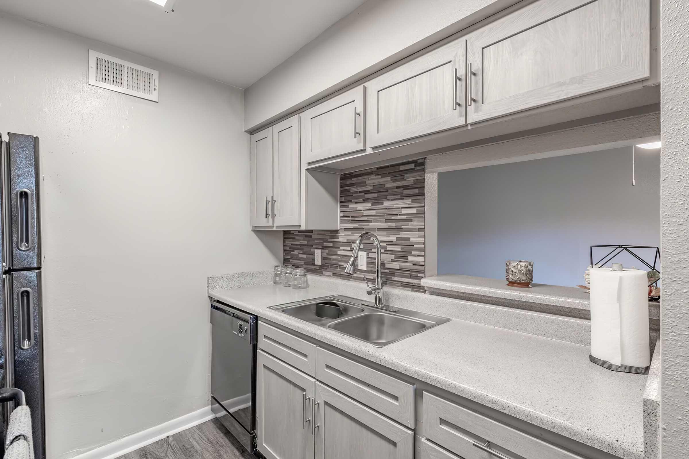 Modern kitchen featuring light gray cabinets, a double sink with a sleek faucet, and a countertop made of light-colored stone. The backsplash is made of small, patterned tiles in shades of gray and brown. Stainless steel appliances include a fridge and dishwasher, adding a contemporary touch to the space.