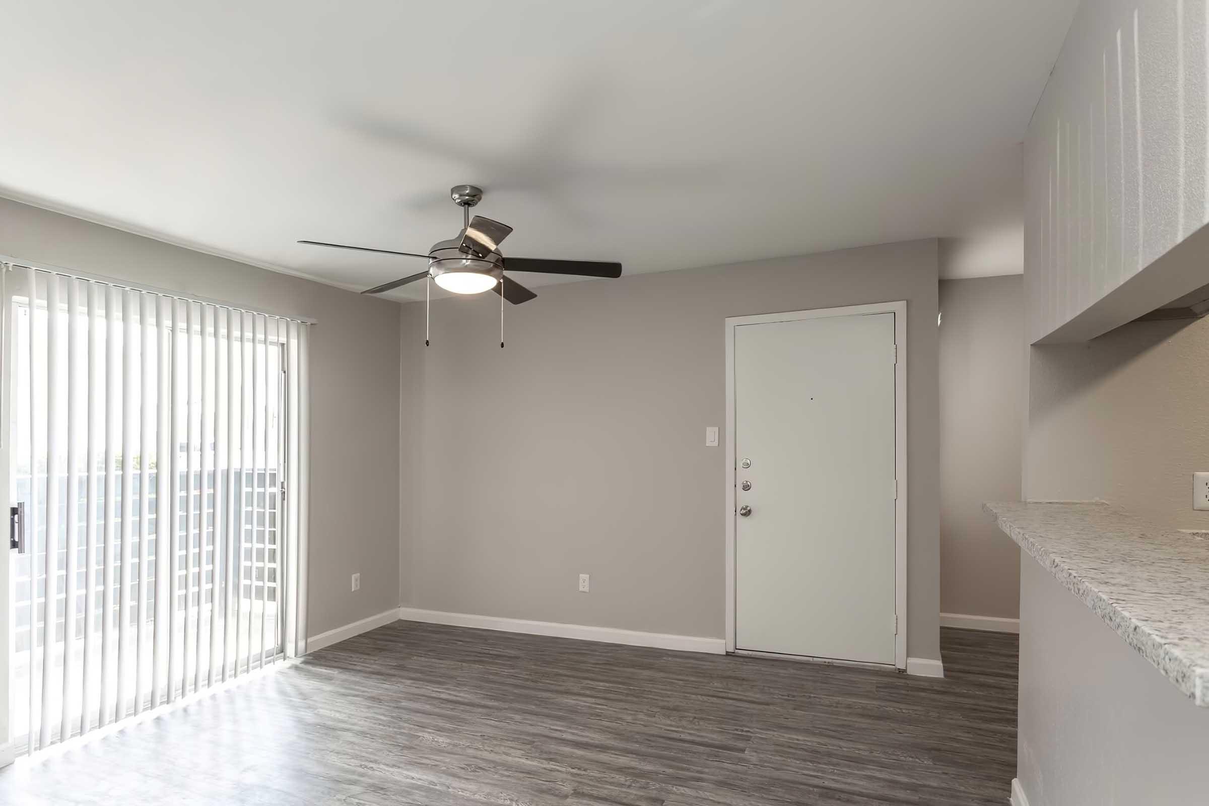 An interior view of a modern living space featuring a ceiling fan, gray walls, and laminate flooring. There is a sliding glass door allowing natural light, along with a closed front door. The adjacent area shows a kitchen counter with a light-colored granite surface.