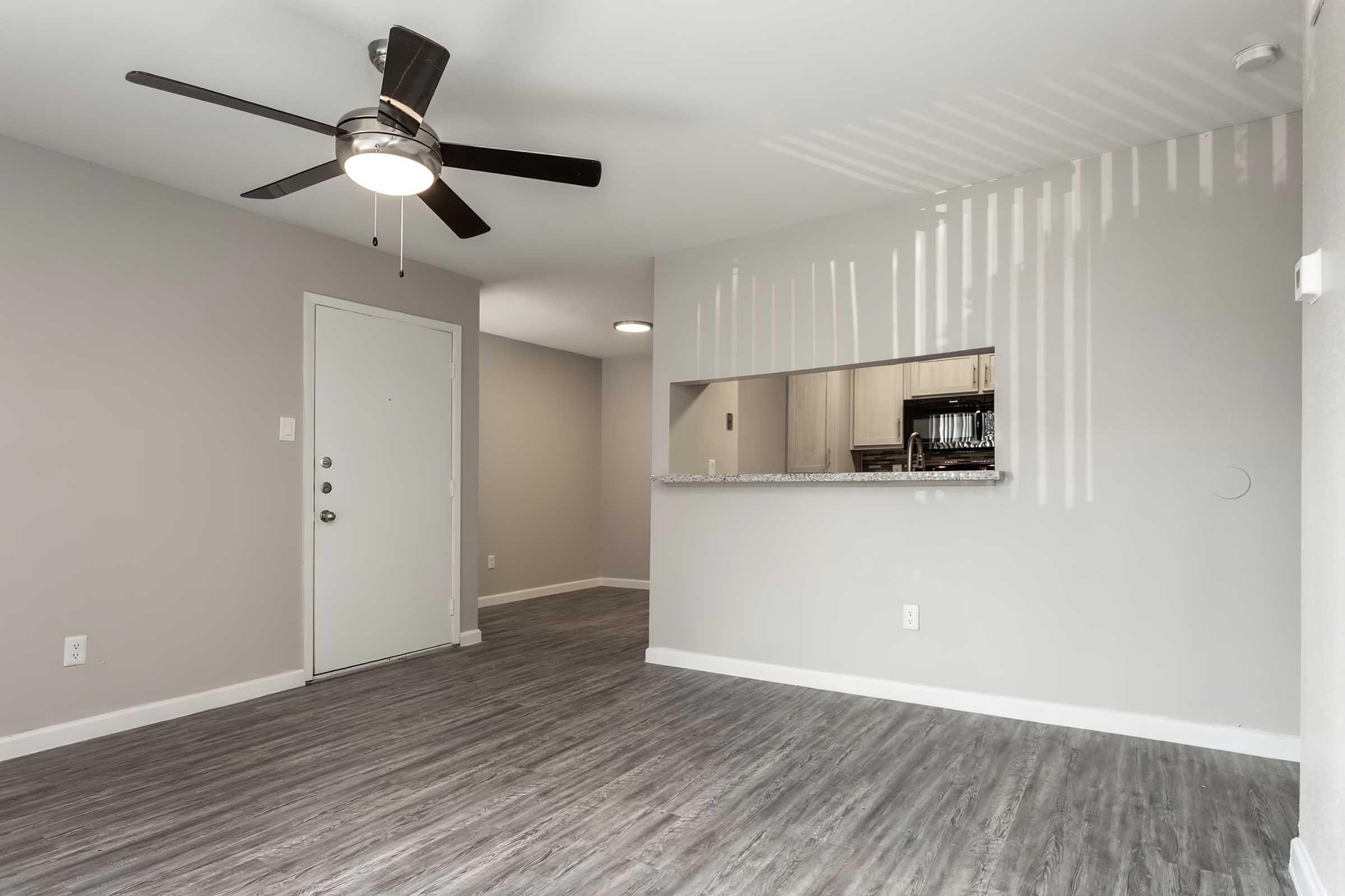 An interior view of a modern living space featuring a light grey wall, a ceiling fan, and a doorway. The floor is covered in a wood-like laminate, and there's a half wall with a counter leading to a kitchen area in the background. Natural light filters in, creating a bright and airy atmosphere.