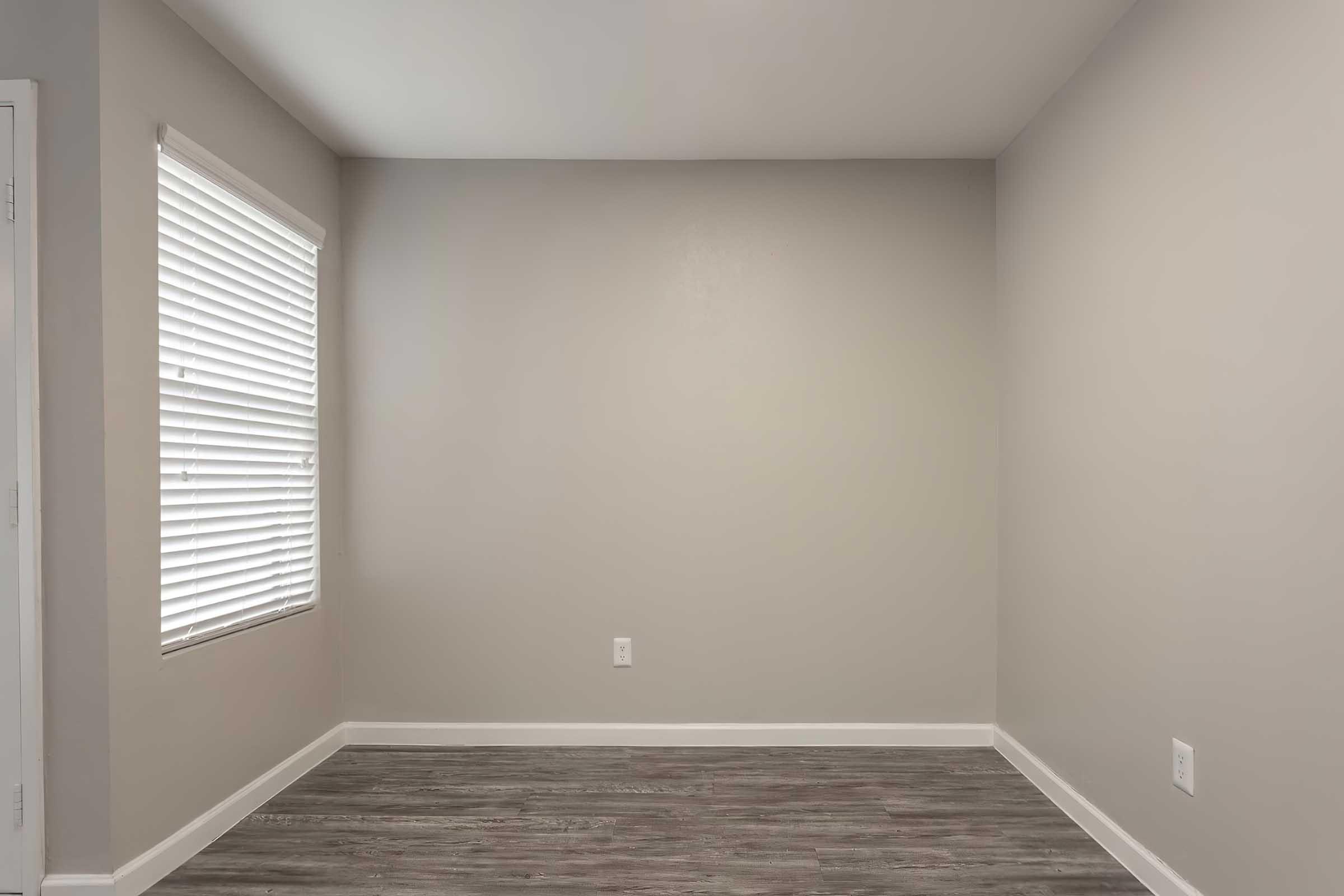 Empty room with light gray walls and a window with white blinds. The floor features laminate flooring in shades of gray, and there is a single electrical outlet and a light switch on the wall. The atmosphere is bright and minimalistic, conveying a sense of openness.