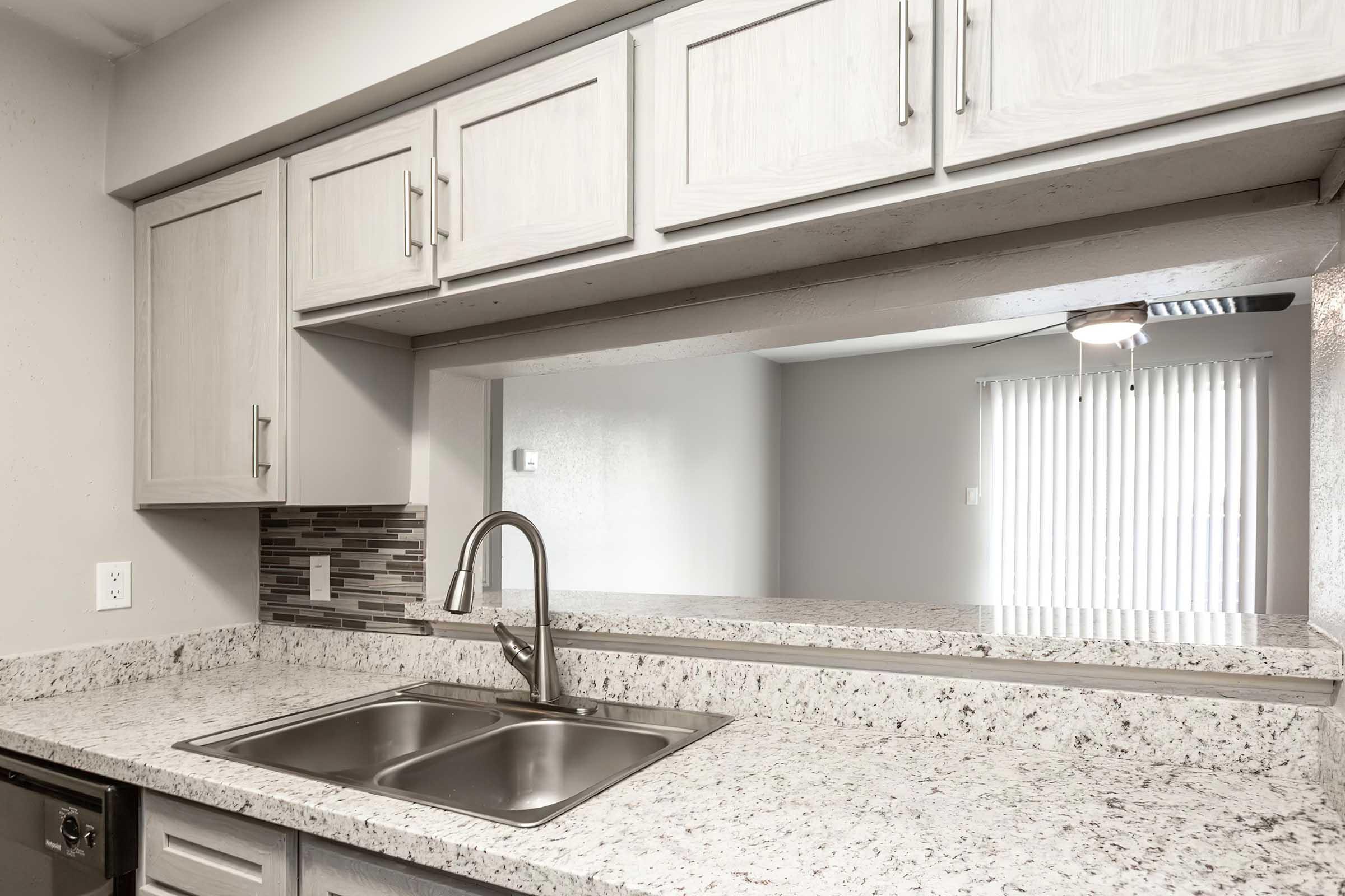 A modern kitchen featuring a double sink set in a granite countertop. Above the sink are light-colored cabinets, and a tiled backsplash adds visual interest. A large window lets in natural light, and the room features neutral-colored walls and blinds. A ceiling light fixture is visible above the sink area.