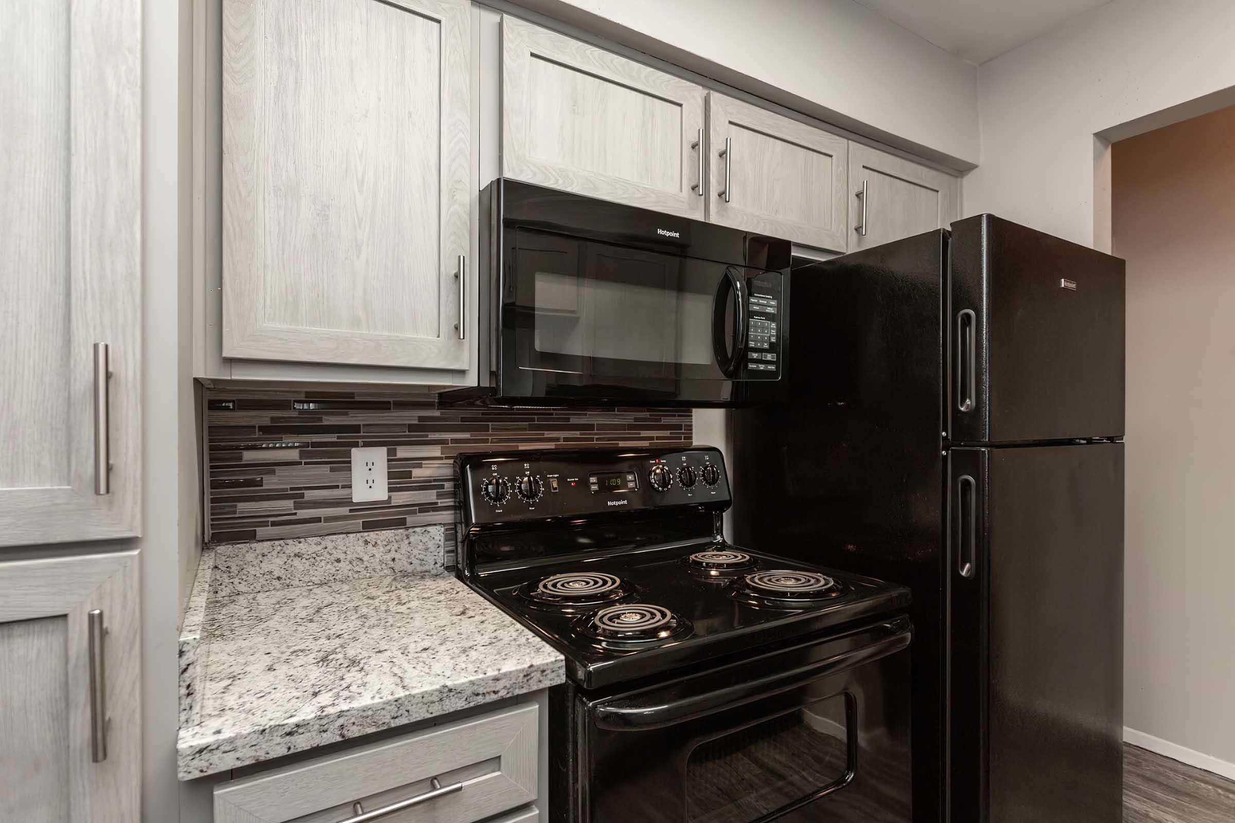 A modern kitchen featuring granite countertops, a black stove, a black microwave above it, and a black refrigerator. The cabinetry is light wood with a textured finish, and the backsplash consists of decorative tiles in various shades. The layout is functional and contemporary.
