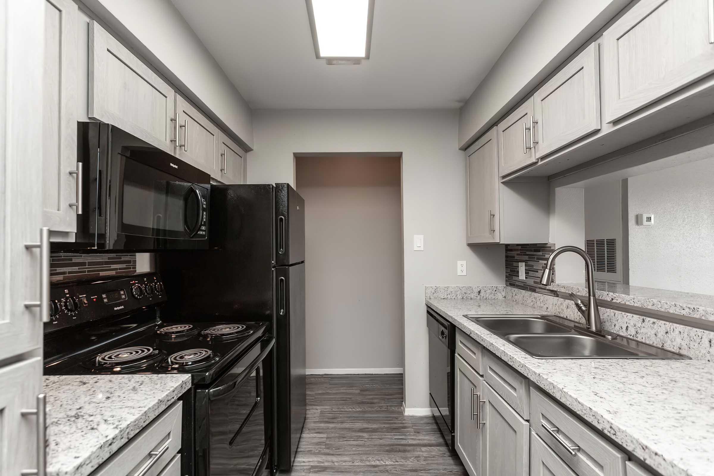 Modern kitchen featuring light gray cabinets, a black refrigerator, oven, and microwave, with a granite countertop. The room has a neutral wall color, and there is a doorway leading to another space. Warm lighting highlights the sleek design and contemporary appliances.