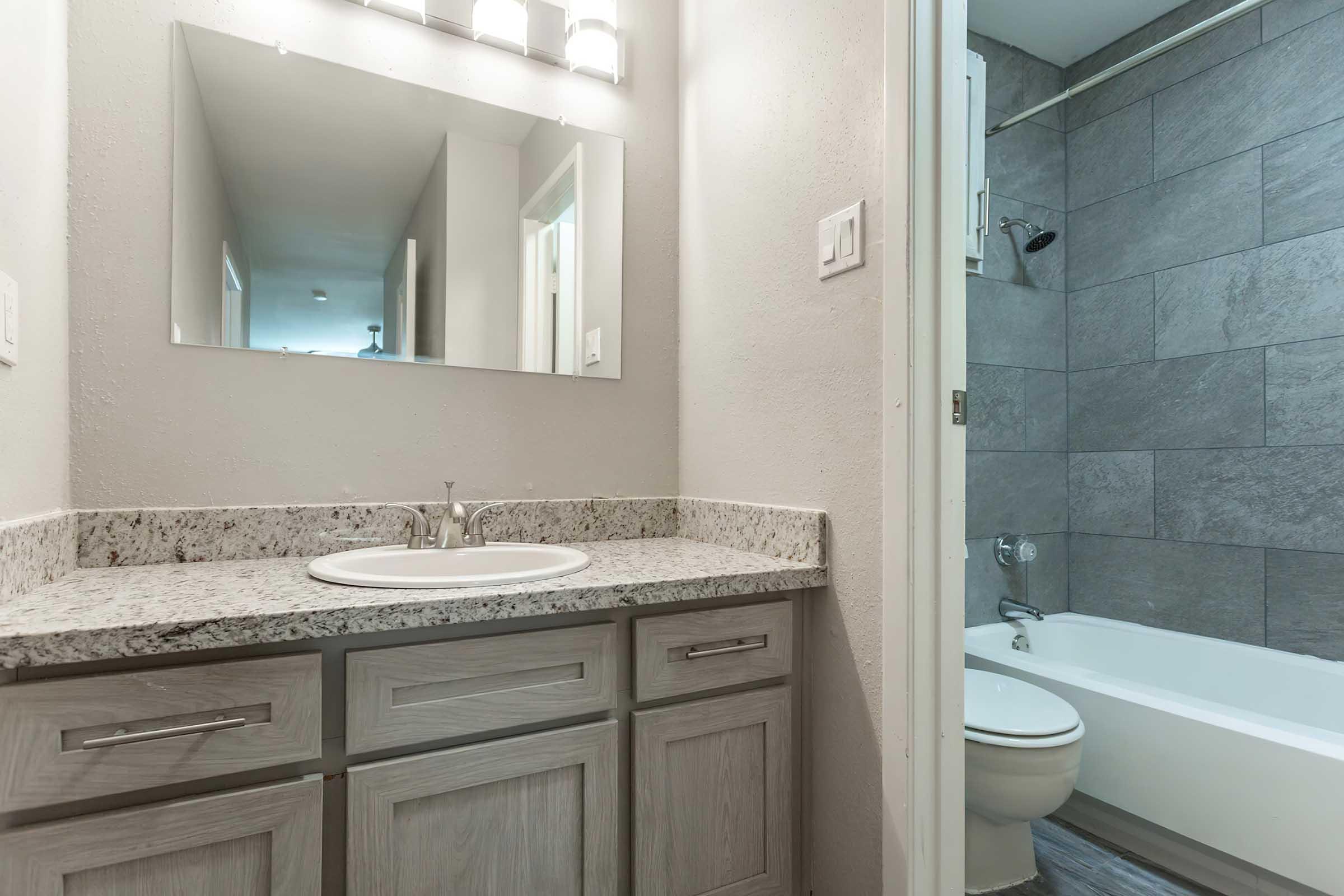 A modern bathroom featuring a granite countertop with a sink, wooden cabinetry, a large mirror above the sink, and a shower area. The bathroom has gray tiled walls and a white bathtub with a showerhead, creating a clean and inviting atmosphere. Natural light is visible in the background.